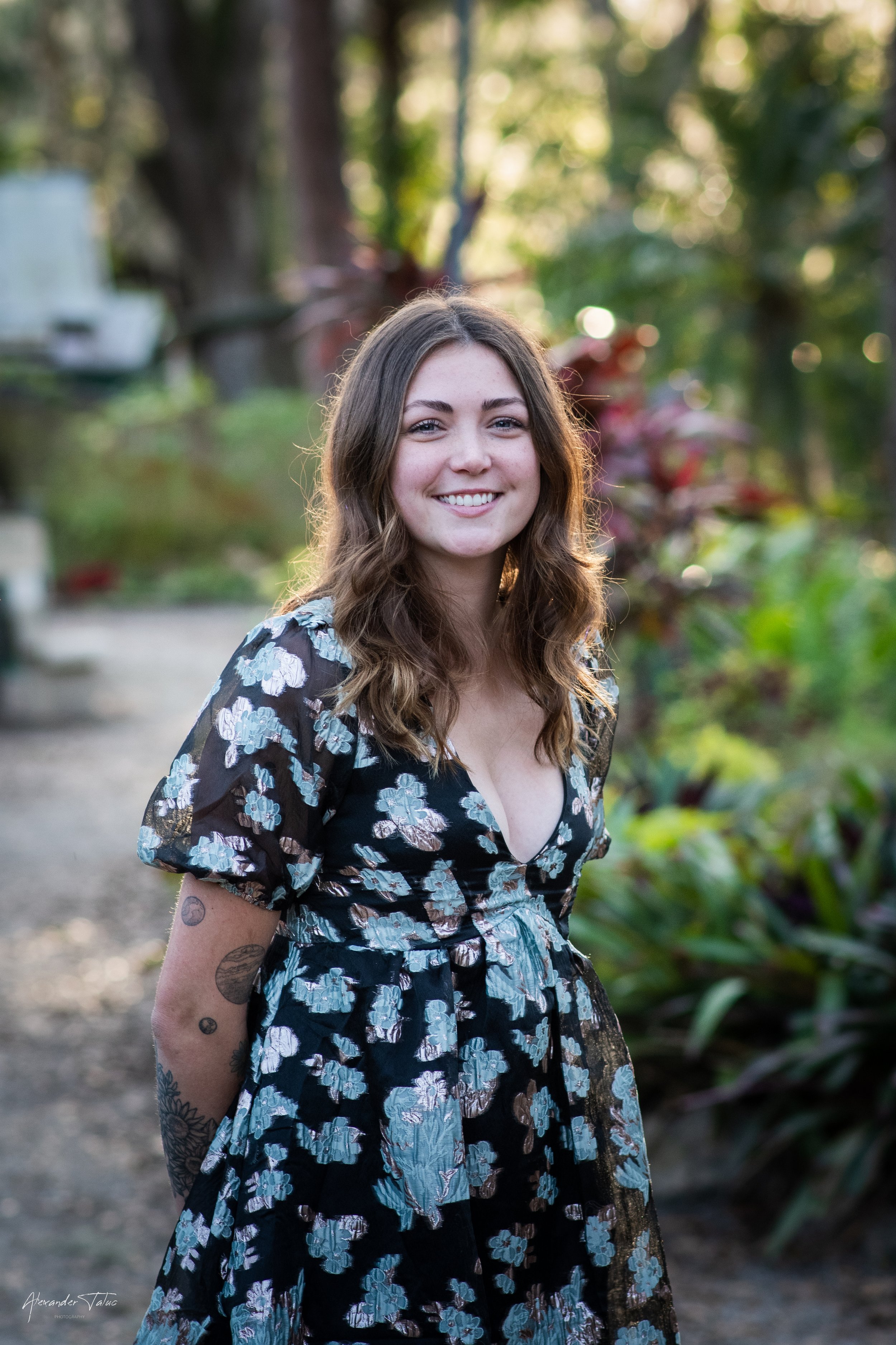 A young woman with wavy brown hair and light skin smiling outdoors during sunset. She is wearing a black floral dress with puffed sleeves, and she has tattoos on her left arm. The background is blurred with trees and plants.