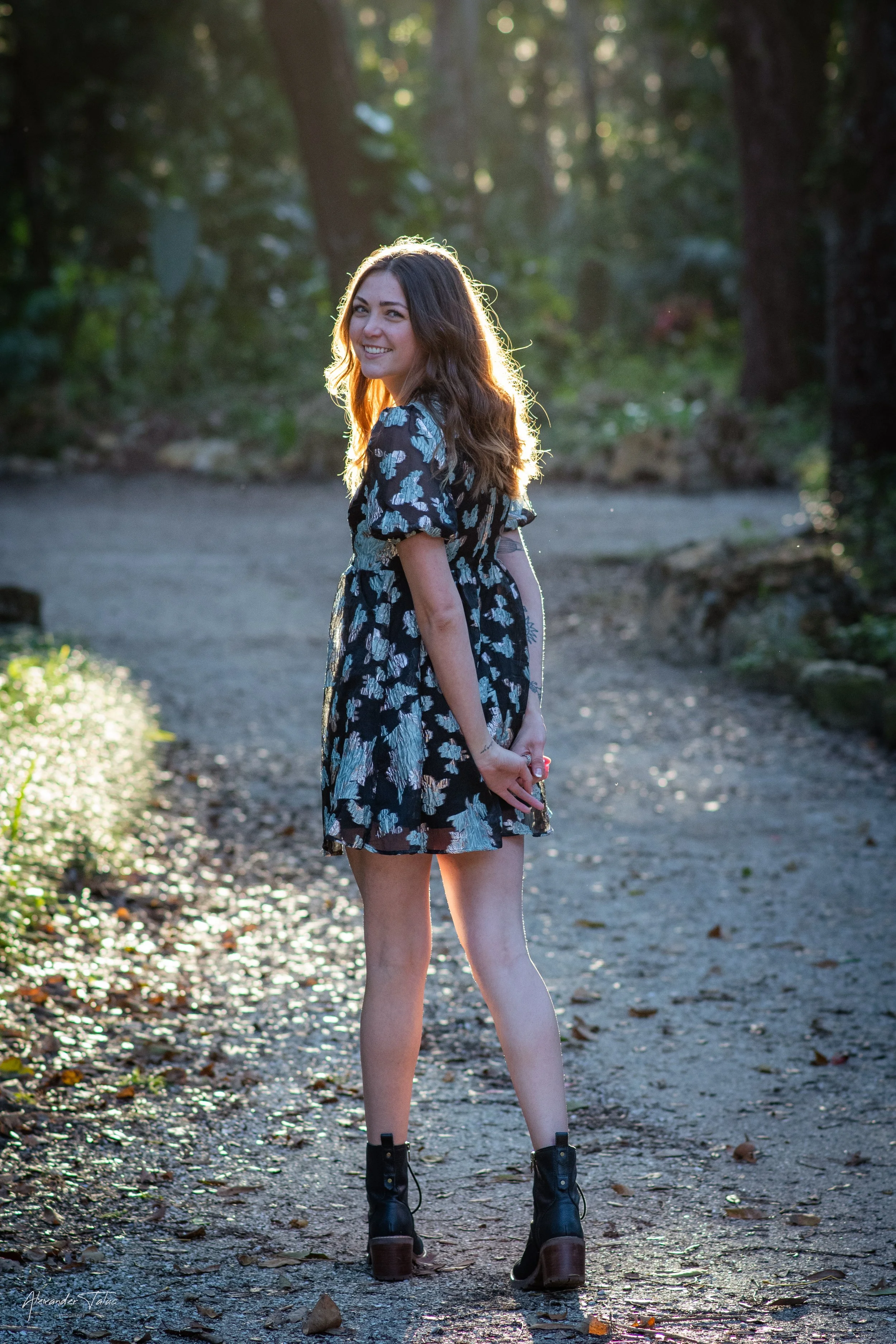 Young woman with long brown hair standing outdoors on a dirt path in a wooded area, smiling, wearing a black floral dress and black boots. Sunlight filters through trees in the background.