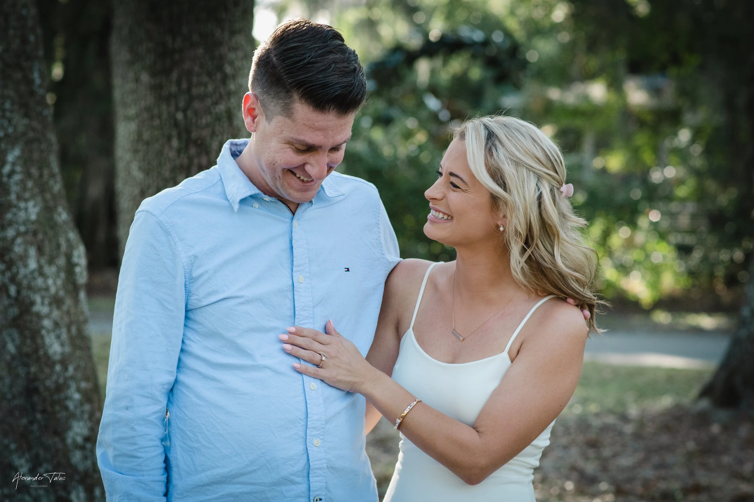 A smiling couple, a man and a woman, standing close together outdoors near a tree with green leaves. The woman has blonde hair, is wearing a white sleeveless dress, and has her hand on the man's chest. The man has dark hair, is wearing a light blue b