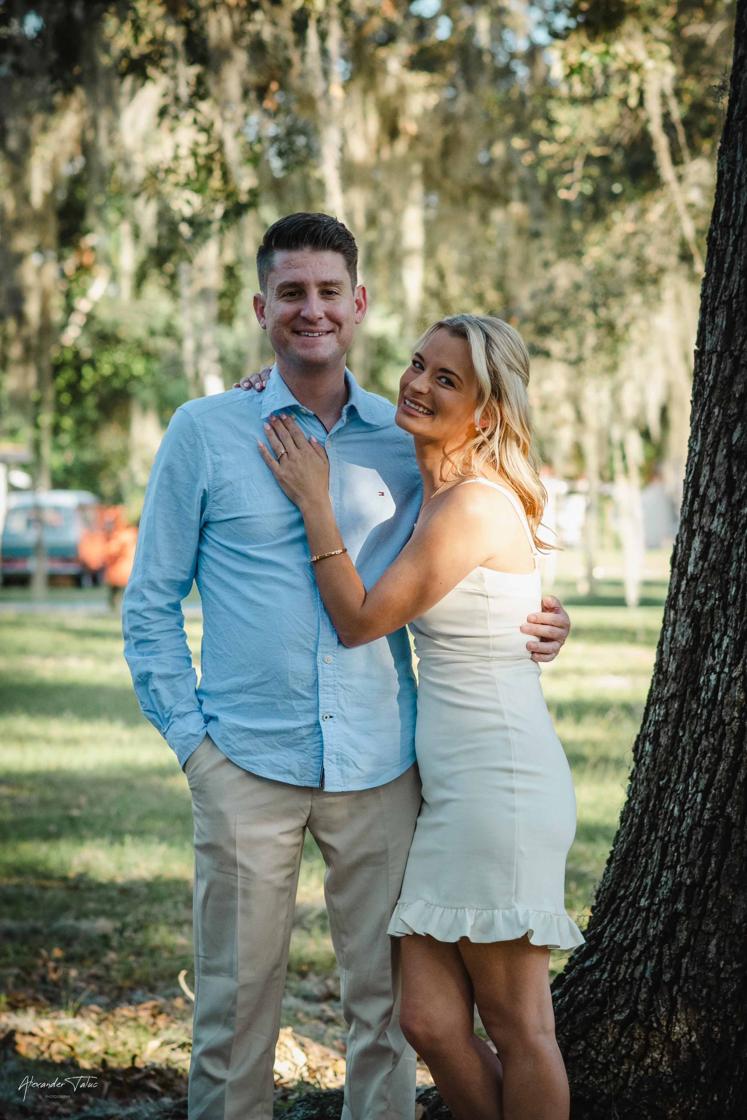 A smiling couple standing close together outdoors in a park with trees in the background. The woman has her hand on the man's chest, wearing a white dress, and the man is dressed in a light blue shirt and beige pants.