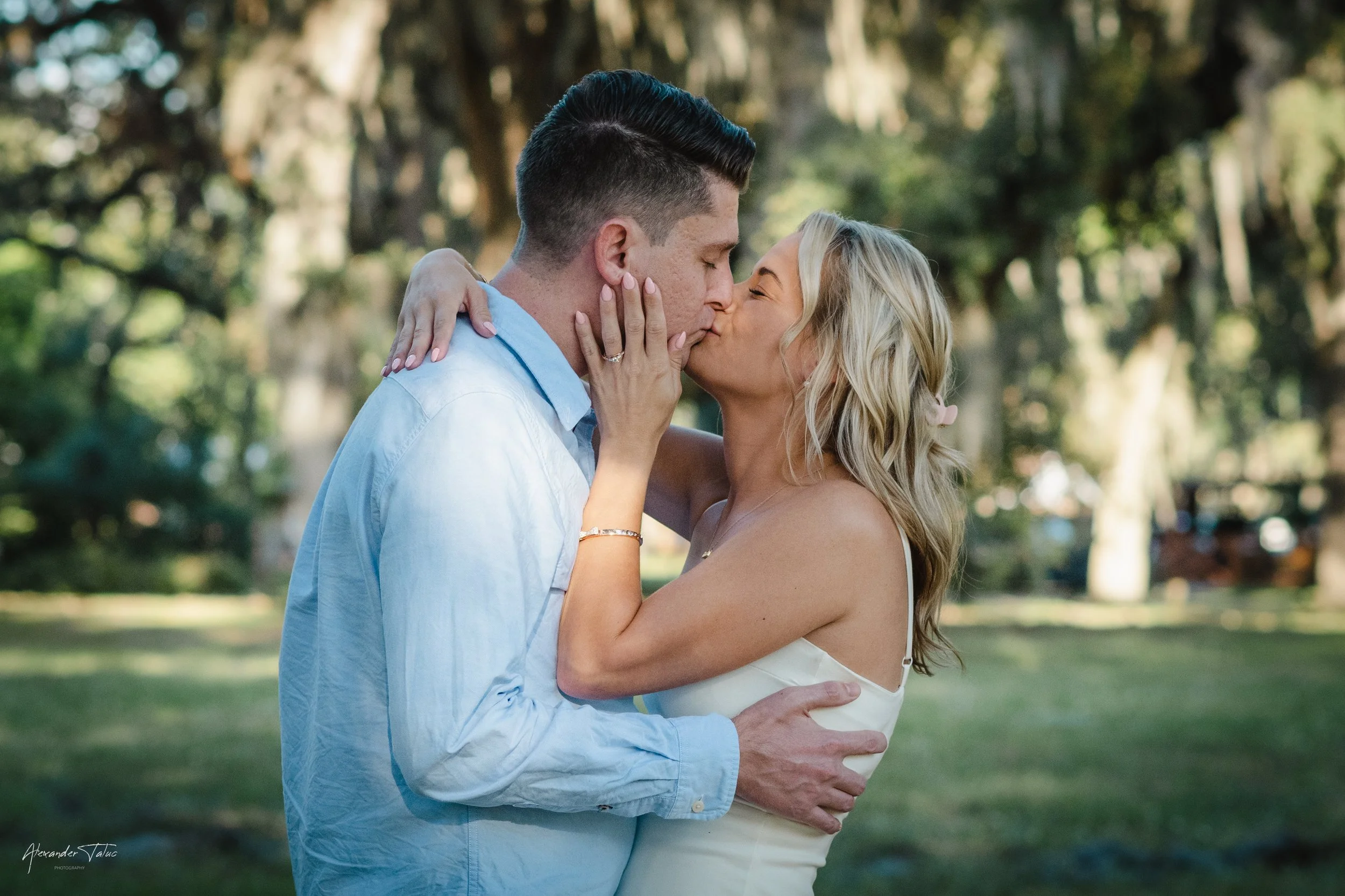 A couple sharing a kiss in a park with trees in the background. The woman is holding the man's face with her left hand, showing an engagement ring. The man has dark hair and is wearing a light blue shirt. The woman has blonde hair and is wearing a wh