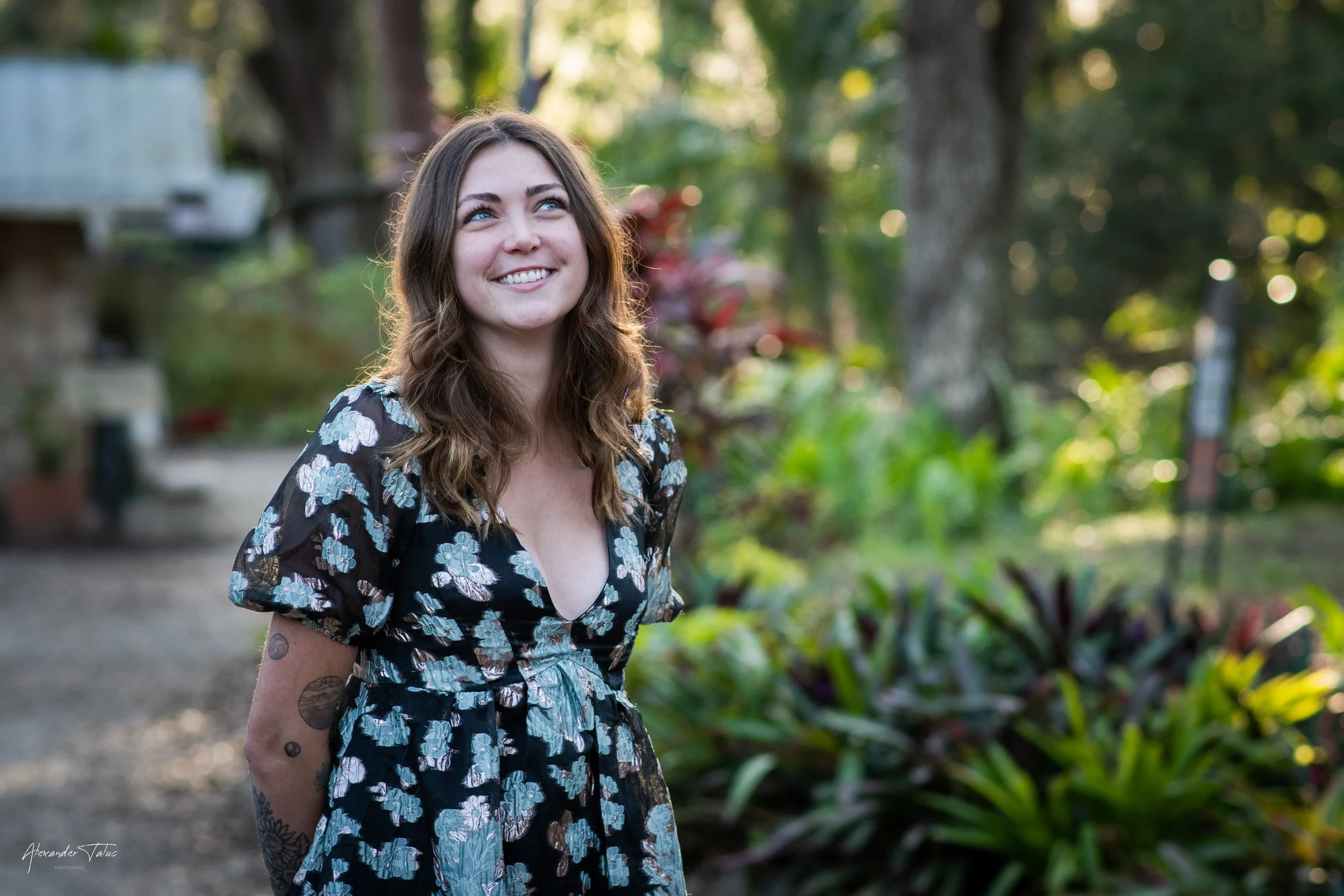 A young woman with long, wavy brown hair and blue eyes, smiling and looking up, standing outdoors in a lush garden with tall green trees and plants, wearing a floral dress with a deep neckline.