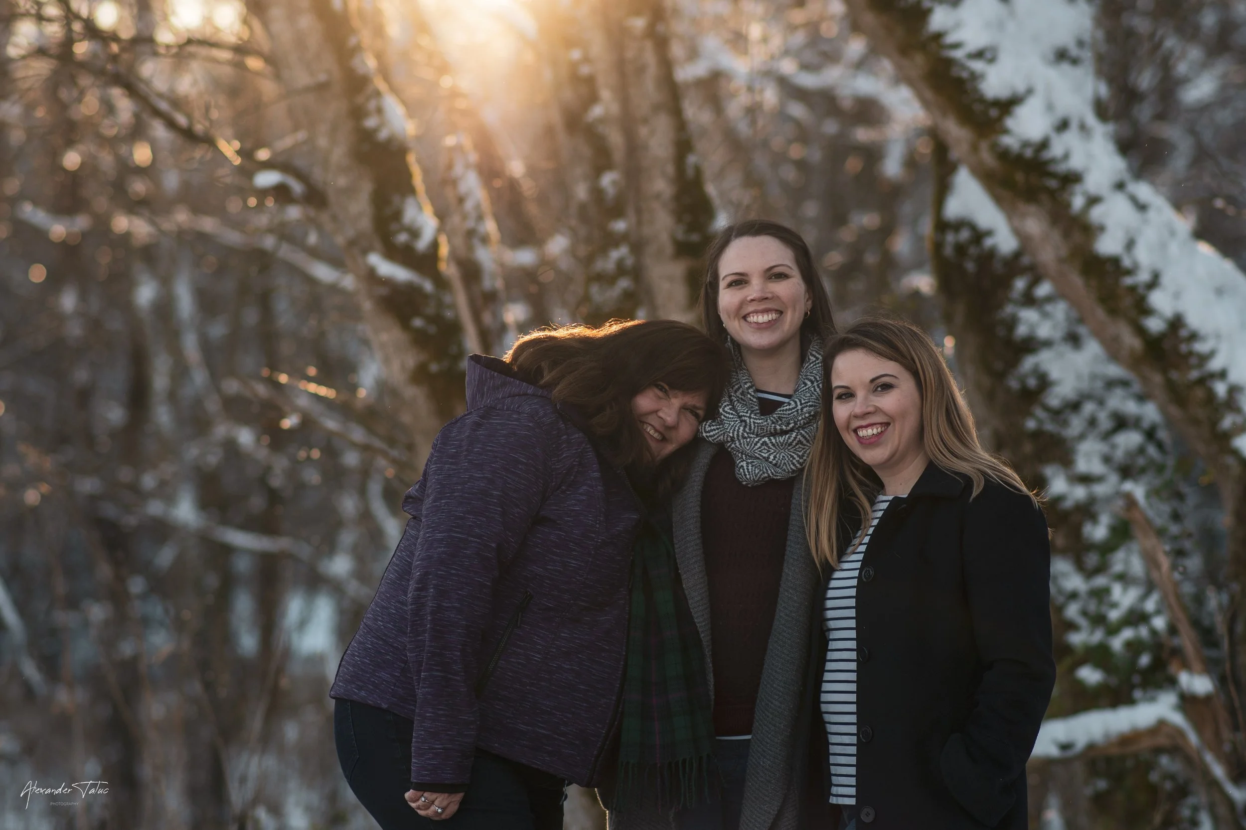 Three women standing close together outdoors in a winter setting with snow-covered trees, smiling at the camera, during sunset.