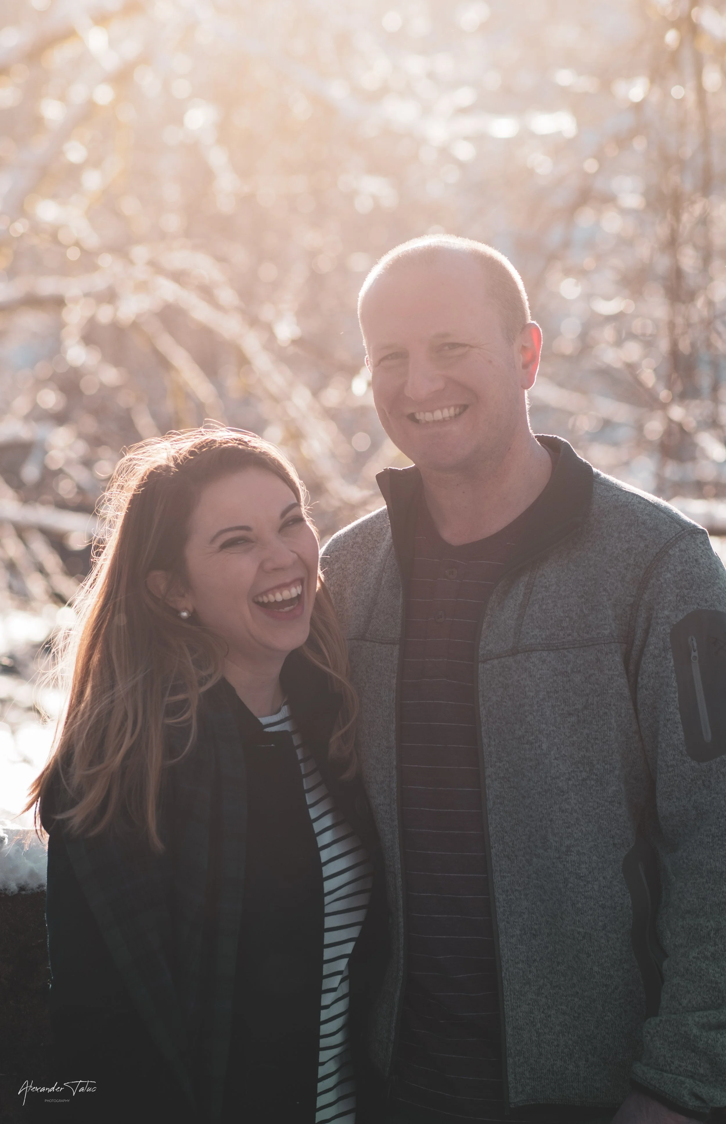 A man and a woman outdoors in a winter setting with snow-covered trees in the background, smiling and enjoying the moment.