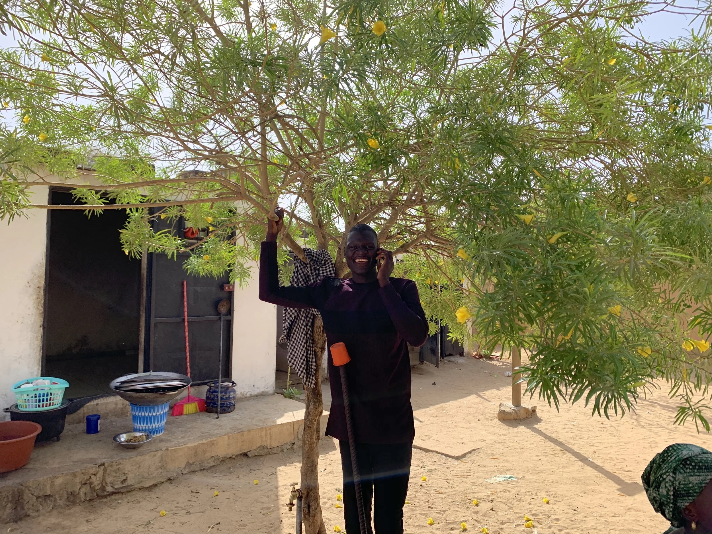 A man smiling and talking on a mobile phone while standing under a tree with yellow flowers in a courtyard. There are household items on a platform, including baskets and bowls, and a building with an open door in the background.
