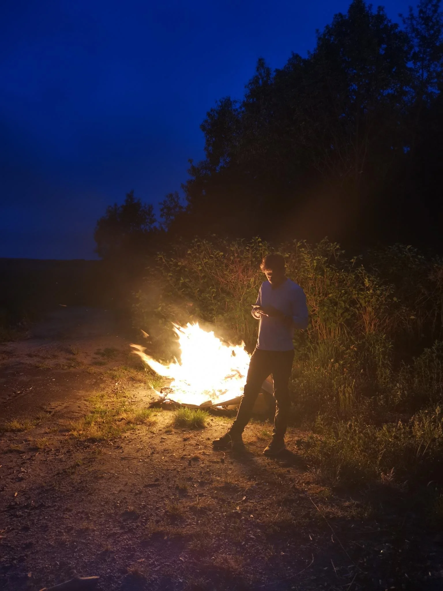 A person standing near a small fire at night with trees and a dark blue sky in the background.