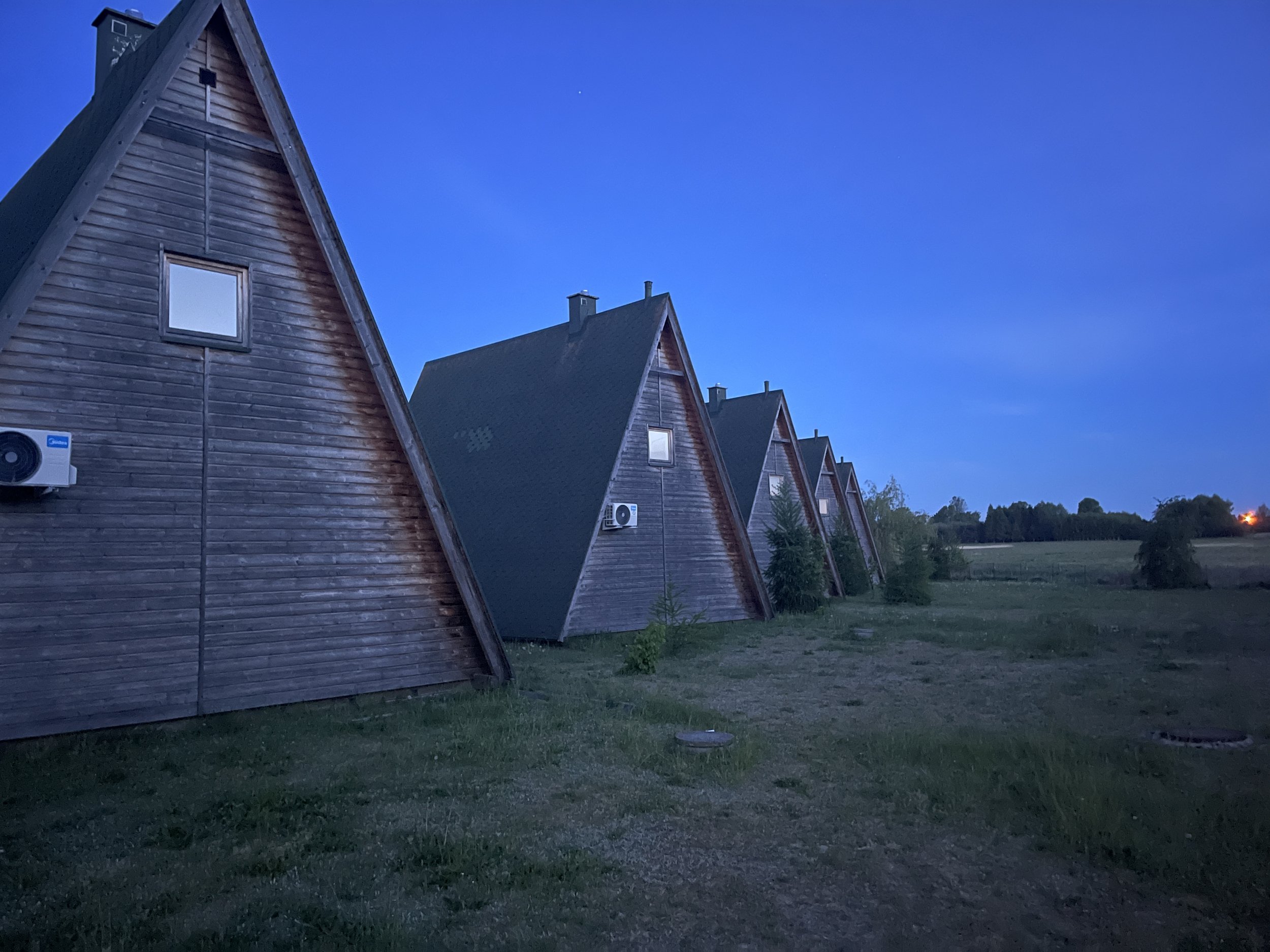 A row of wooden houses with triangular roofs and small square windows at dusk, surrounded by a grassy area and a few trees in the background.