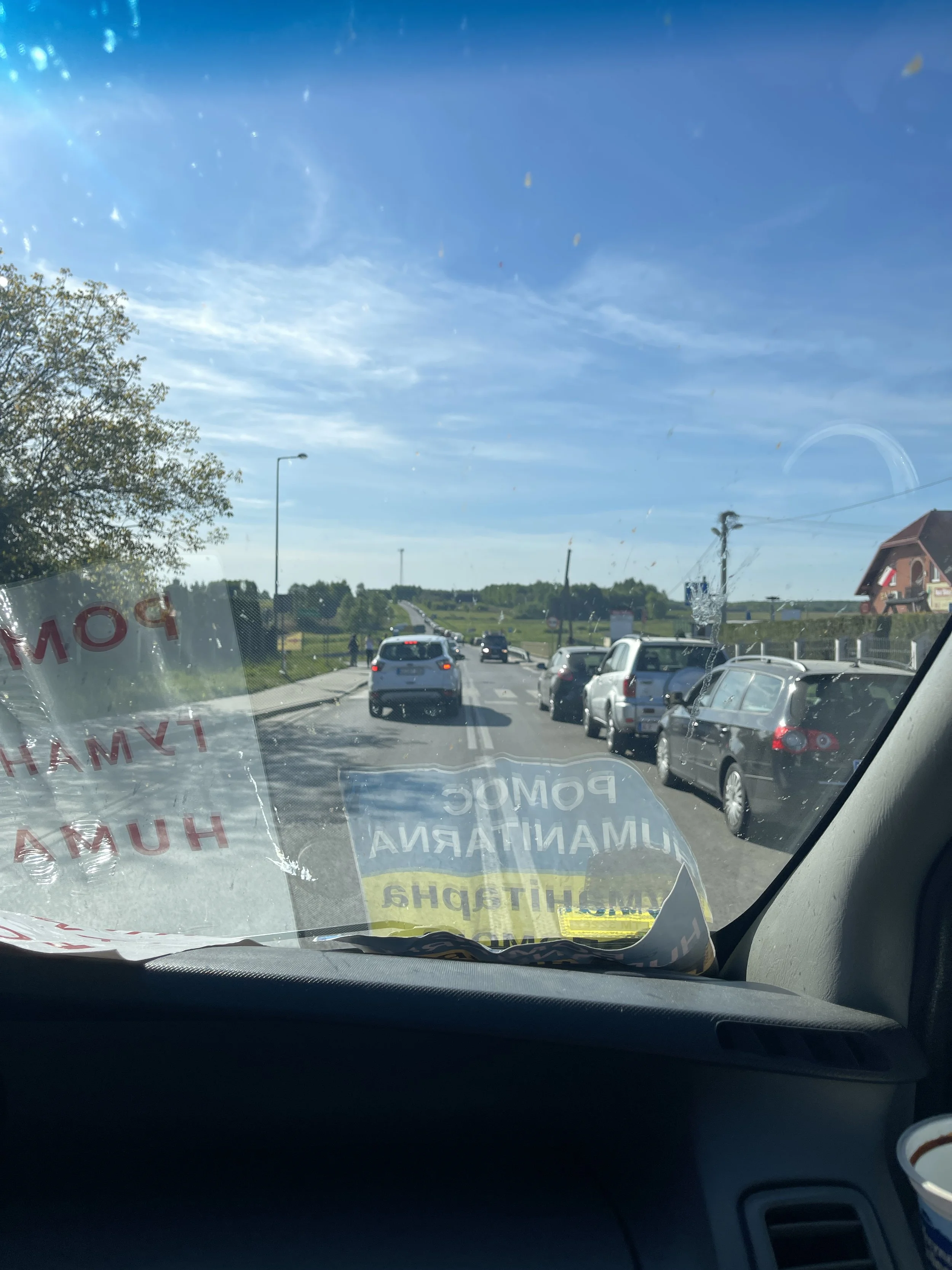 View from inside a vehicle looking out through the windshield at a street scene with cars in traffic, trees, and buildings, with a clear blue sky overhead. Some stickers or signs are partially visible on the lower part of the windshield.