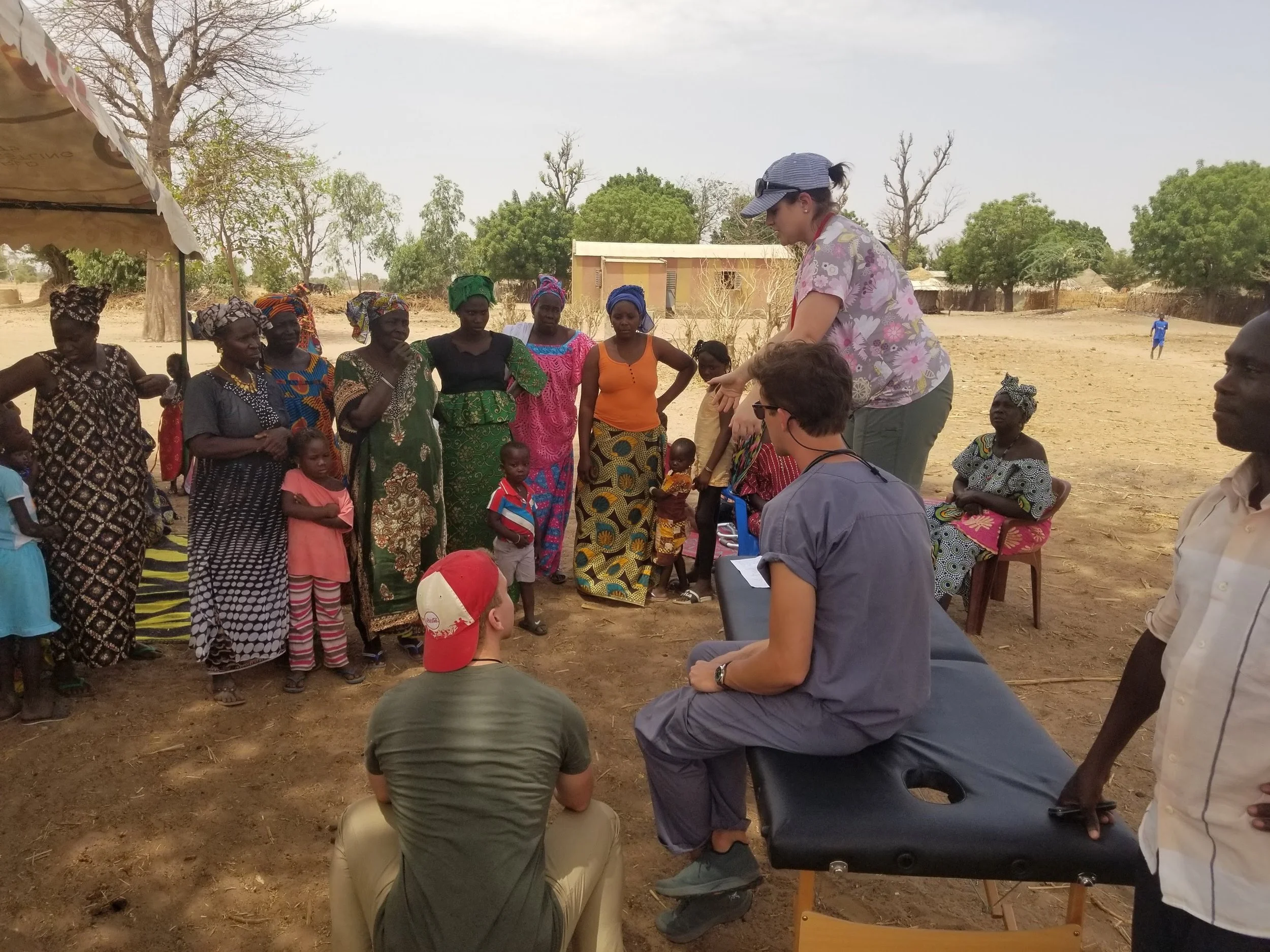 A group of women and children in Africa gathered outdoors on a dirt area, listening to a woman standing and speaking. Some people are seated, and others are standing. The background features trees, a small building, and a partly cloudy sky.