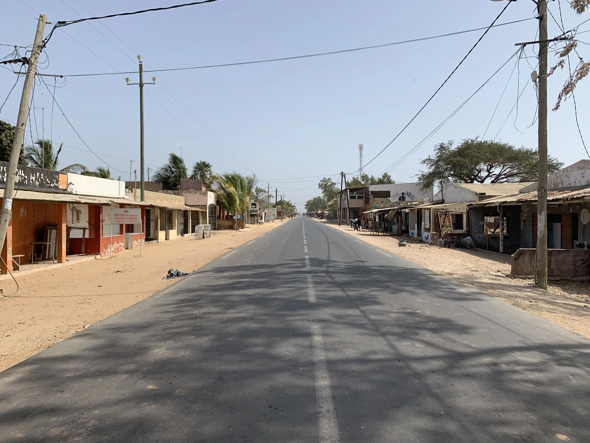 A small town street scene with shops on either side and overhead power lines. The street is mostly empty, with a bicycle and few signs visible. The buildings appear slightly worn, and there are some trees in the background.