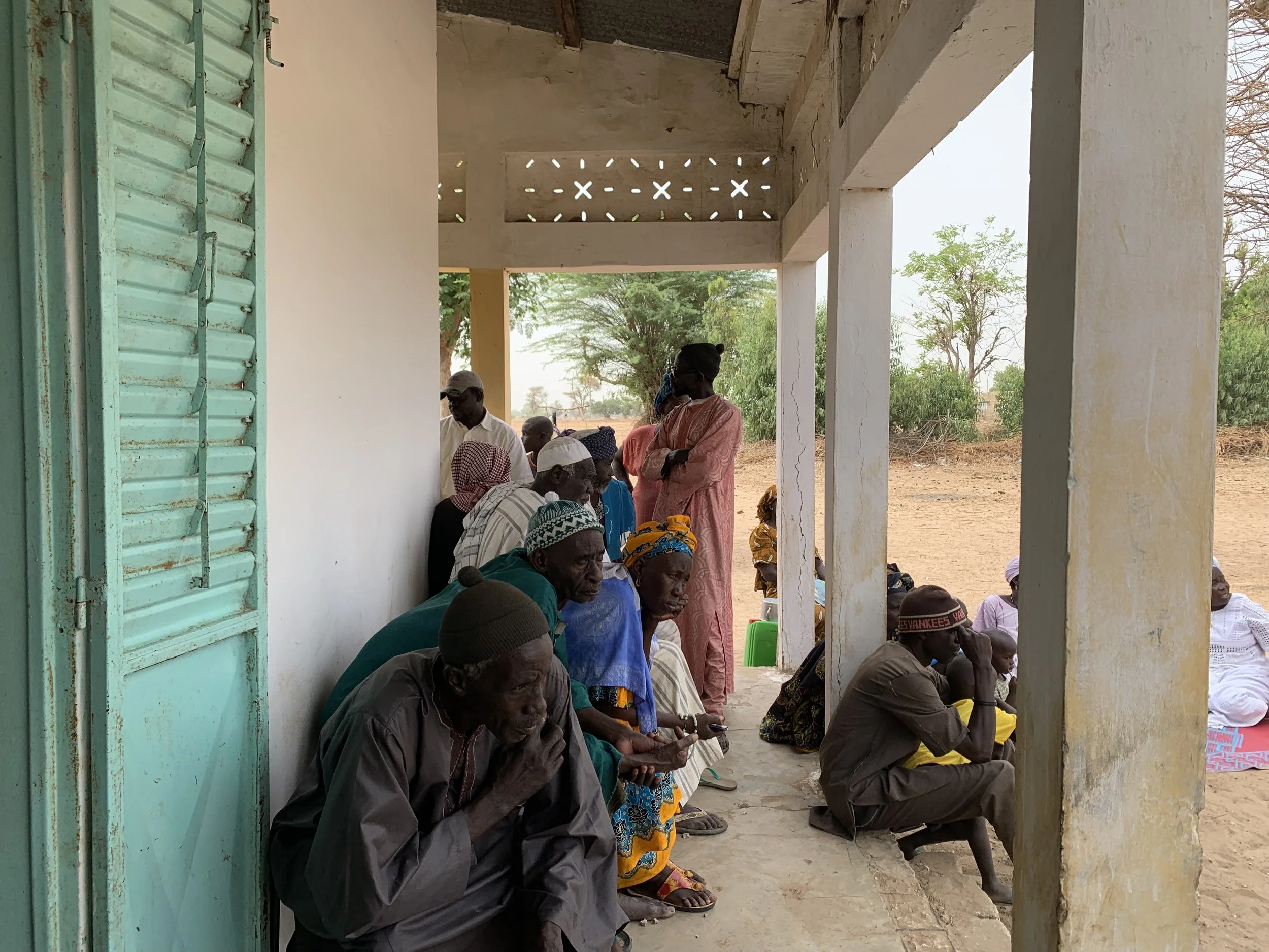 A group of people, some sitting and some standing, gathered under a covered outdoor area with a dirt ground and trees in the background, likely in a rural setting.