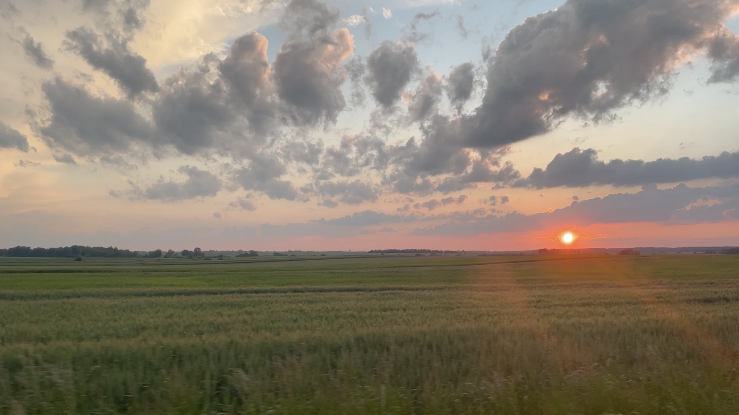 A landscape of green fields with a partly cloudy sky at sunset. The sun is near the horizon, emitting a warm glow and pinkish hues in the clouds.