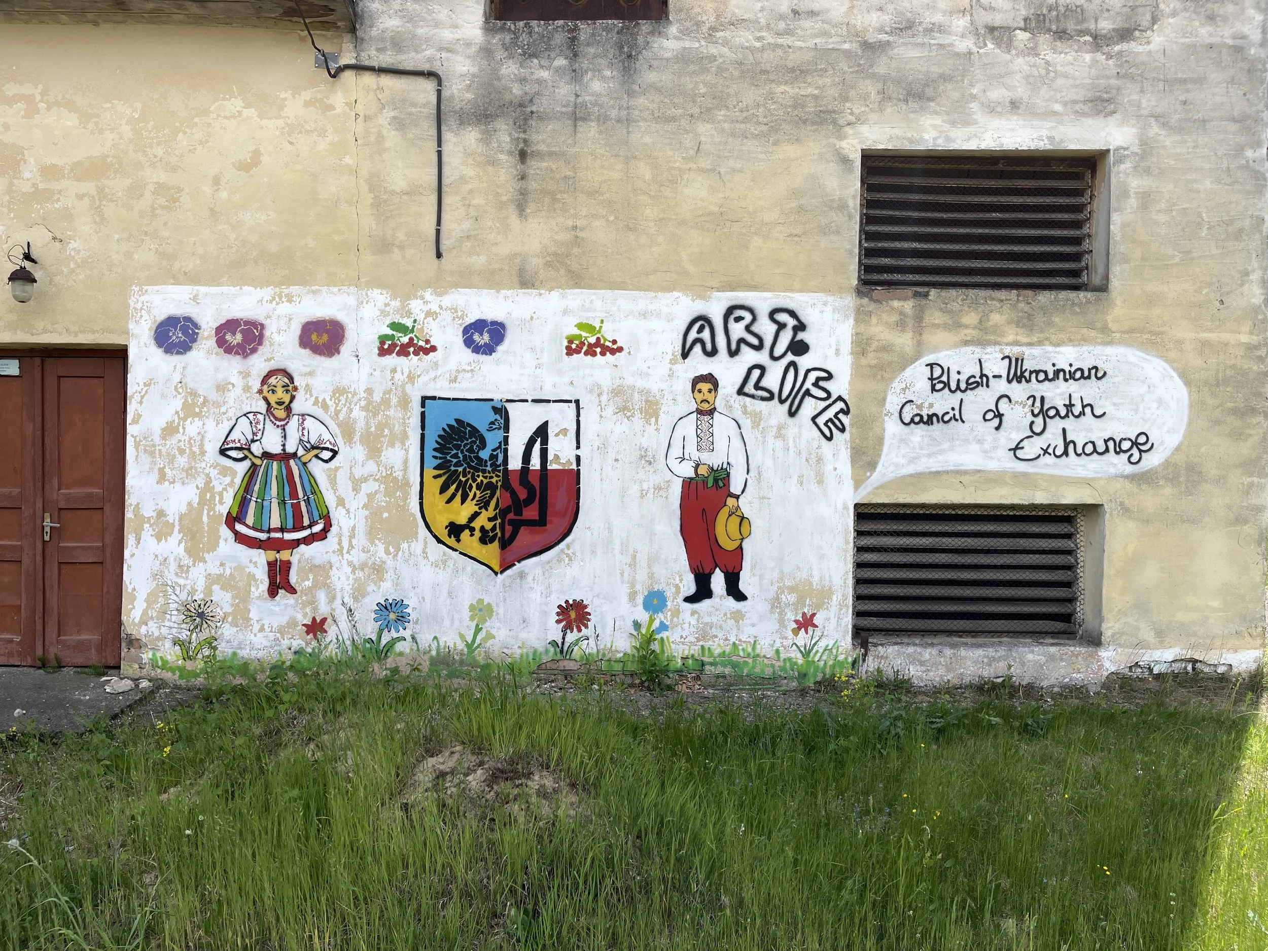 Muralon a weathered beige wall featuring traditional Ukrainian artwork, with two characters dressed in Ukrainian folk costumes, a woman in a floral dress and a man in embroidered attire, surrounded by colorful flowers and a shield emblem. A speech bubble states, "Blish-Ukrainian Council of Yath Exchange."