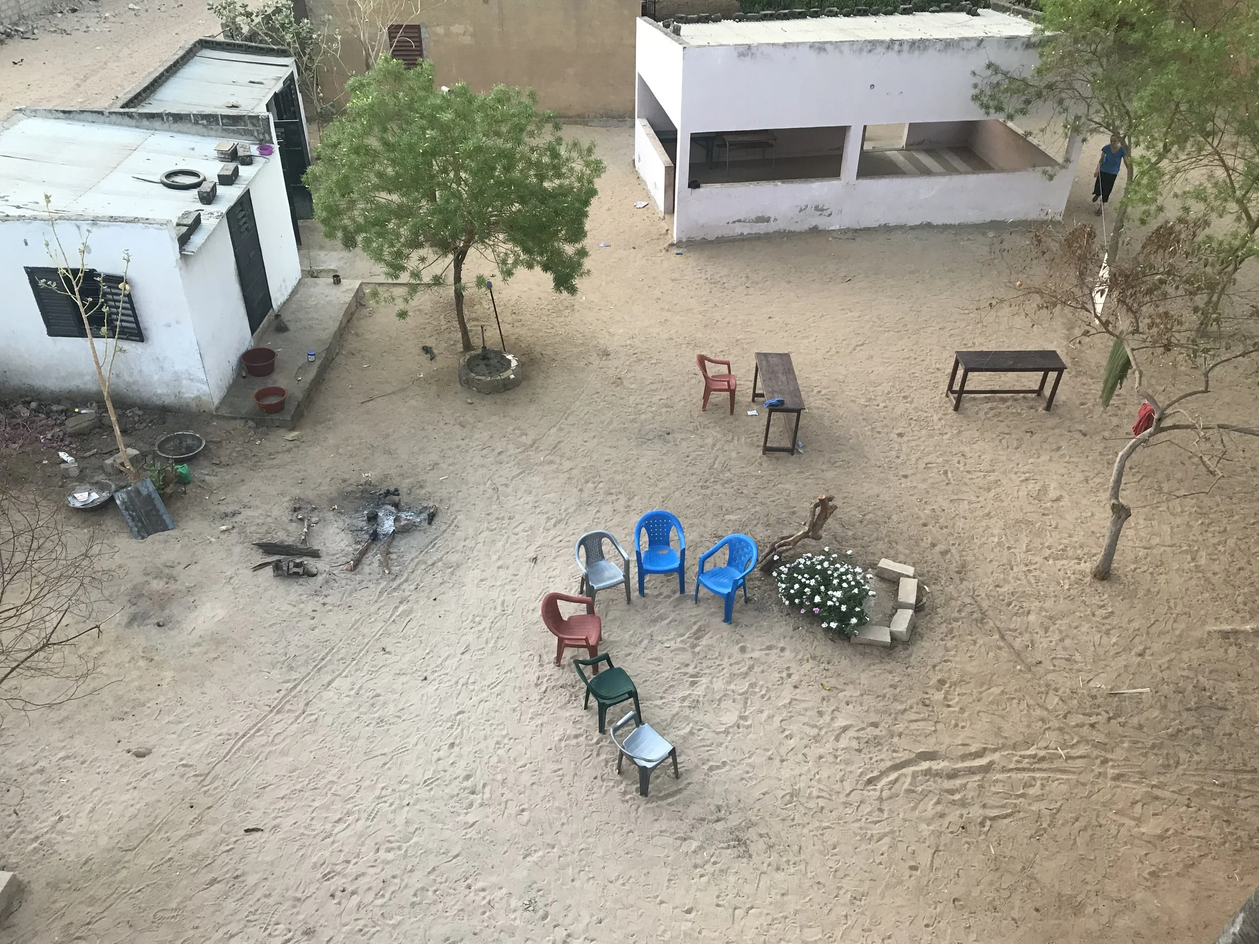 A dirt yard with several plastic chairs arranged in a circle, a table, trees, and a small white building. There are some scattered objects and debris on the ground, and a woman walking in the background.