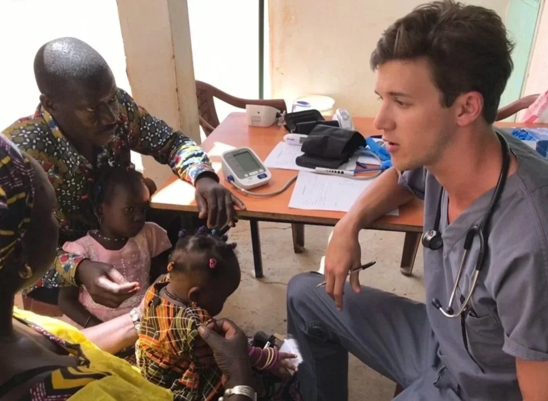 A young handsome man healthcare worker with a stethoscope around his neck consulting with a group of African women and children in a clinic setting, with medical equipment and paperwork on a table in the background.