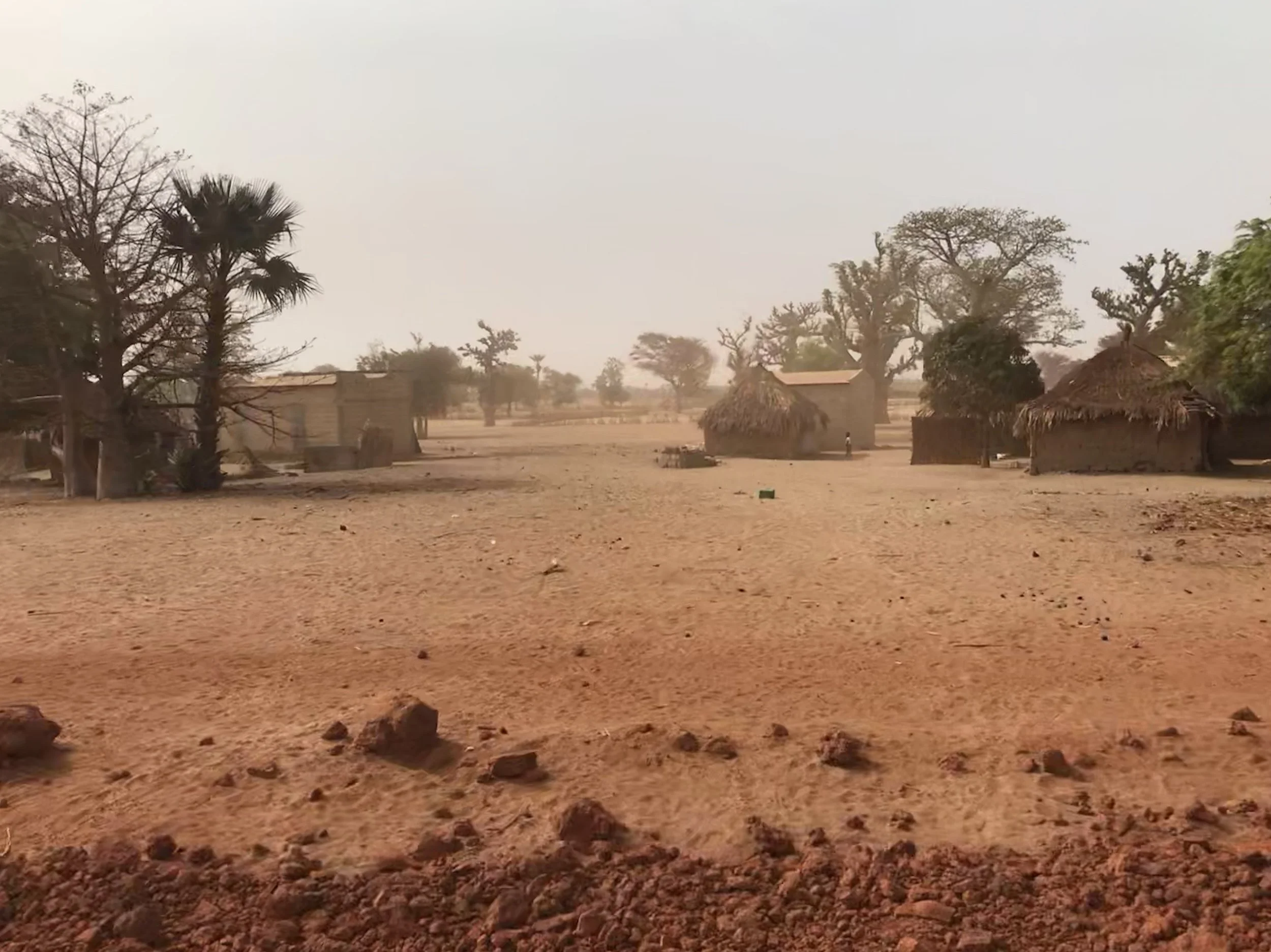 A rural village scene with traditional huts, trees, and dry, cracked ground in a dusty environment.