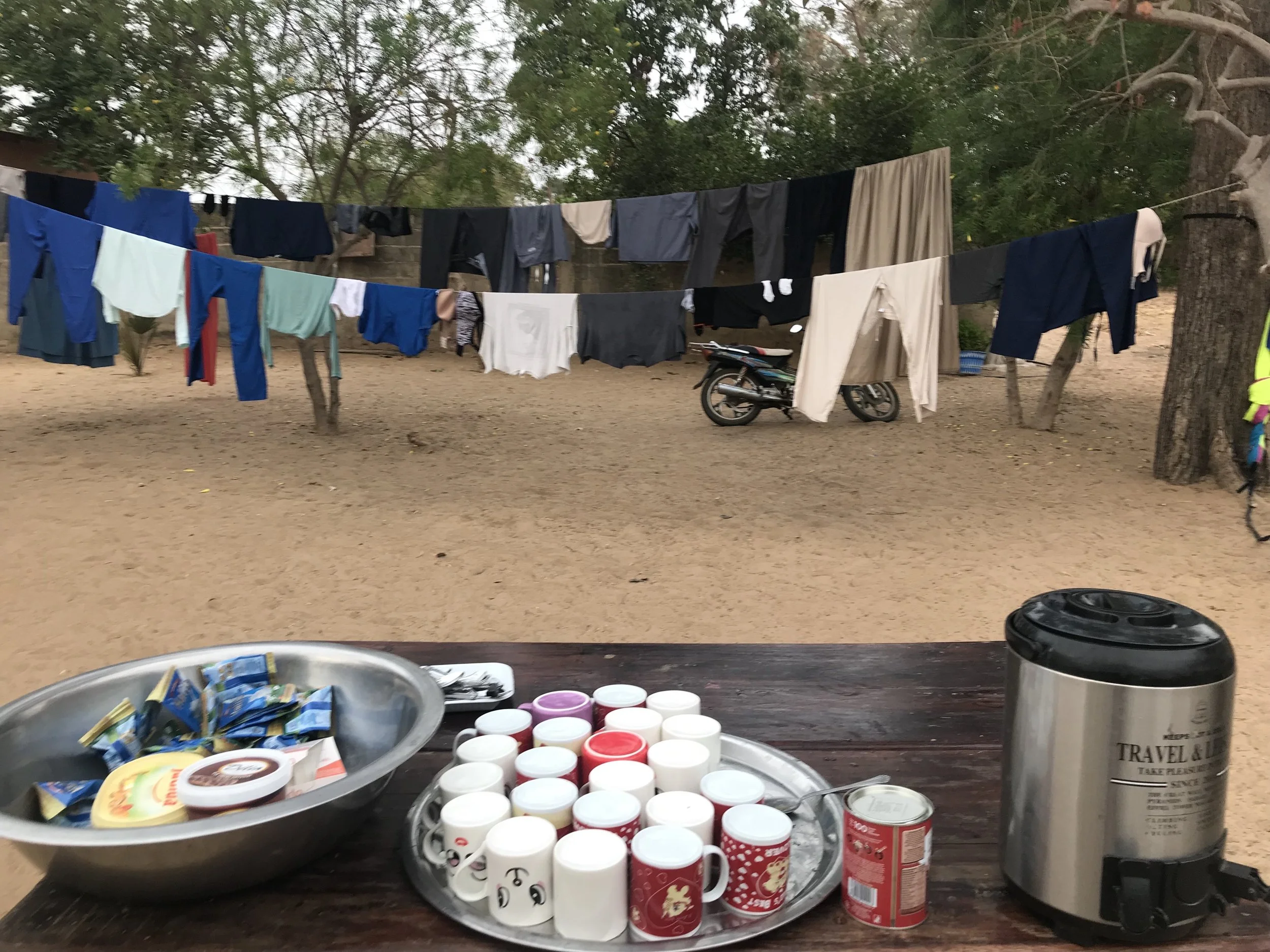 Table with various food items including cups, containers, and snack packaging, with clothes hanging on a line in the background and trees surrounding an outdoor area.
