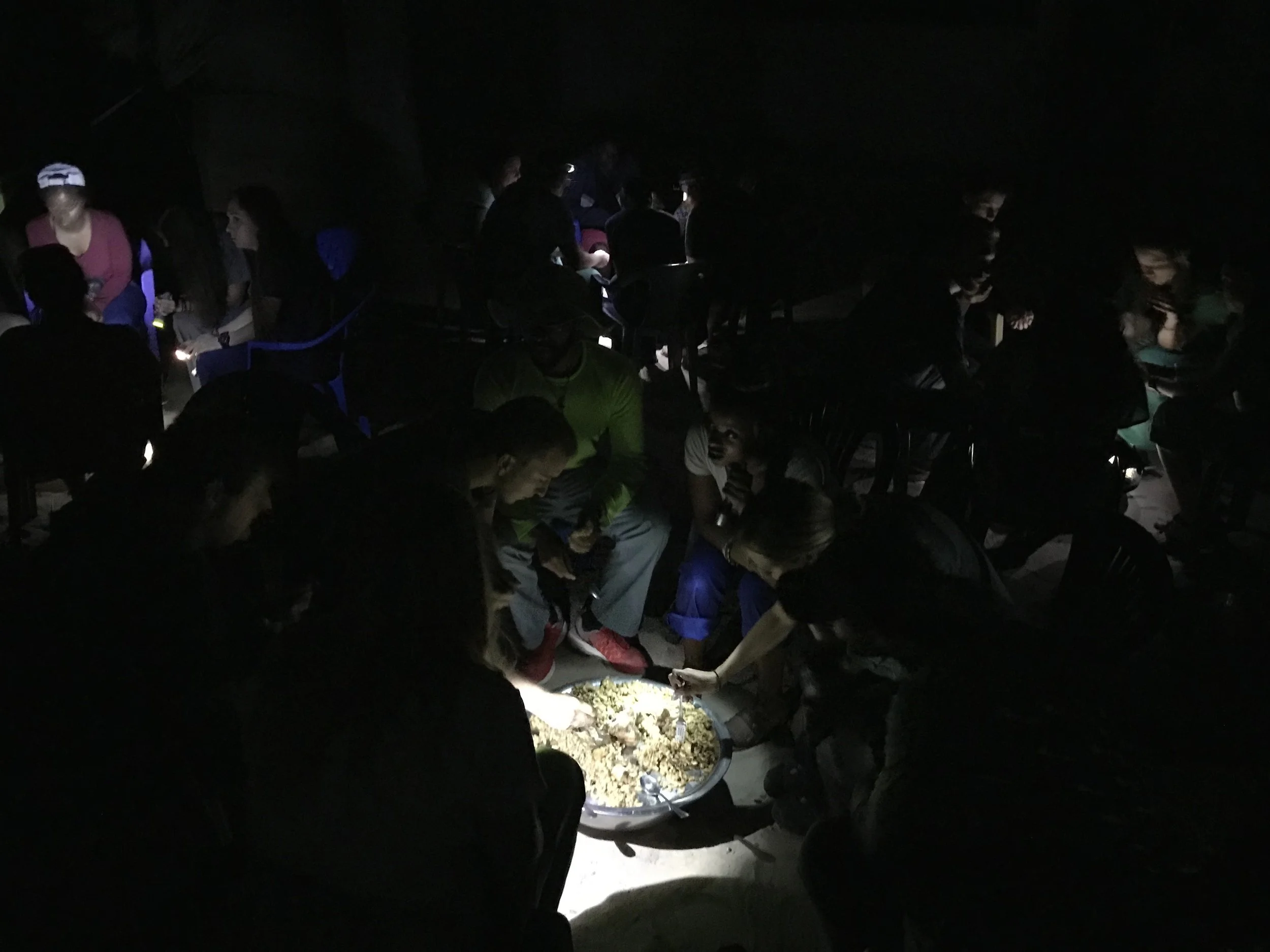 People gathered around a large dish of food under low lighting at night.