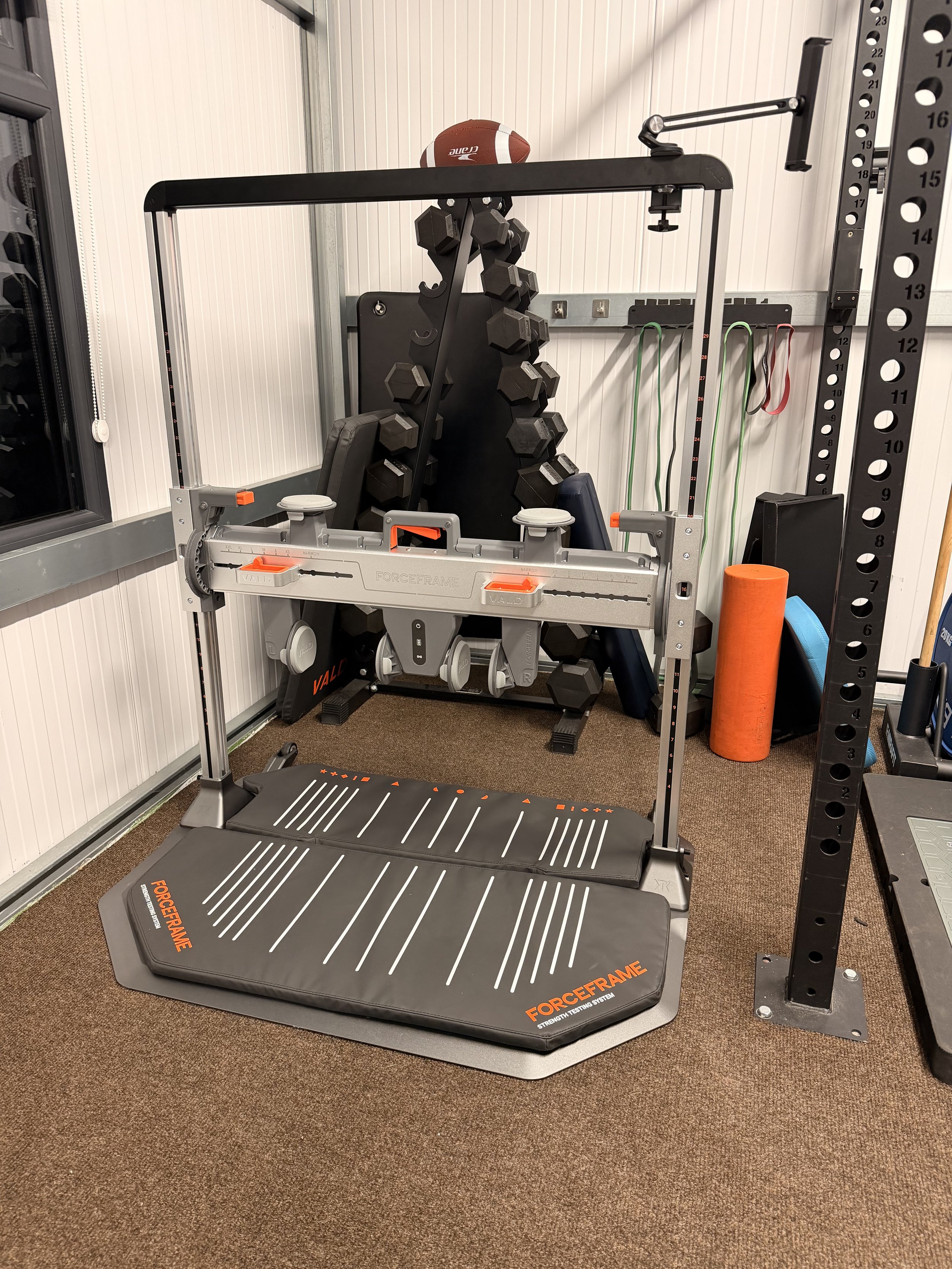 VALD Forceframe testing system, a set of dumbbells on a rack, exercise resistance bands hanging on the wall, and a foam roller.