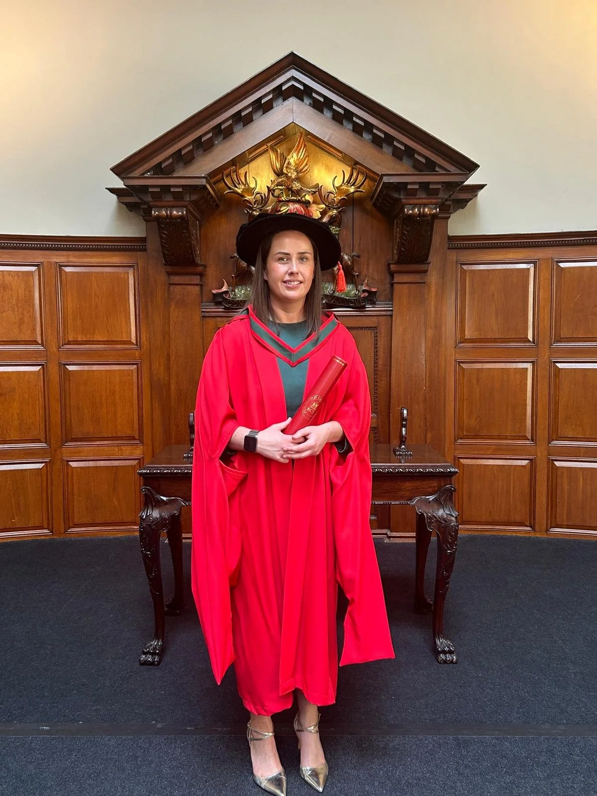 Clinic owner Fiona in a red graduation gown and cap holding a PhD diploma in a room with wood paneled walls and ornate wooden furniture.