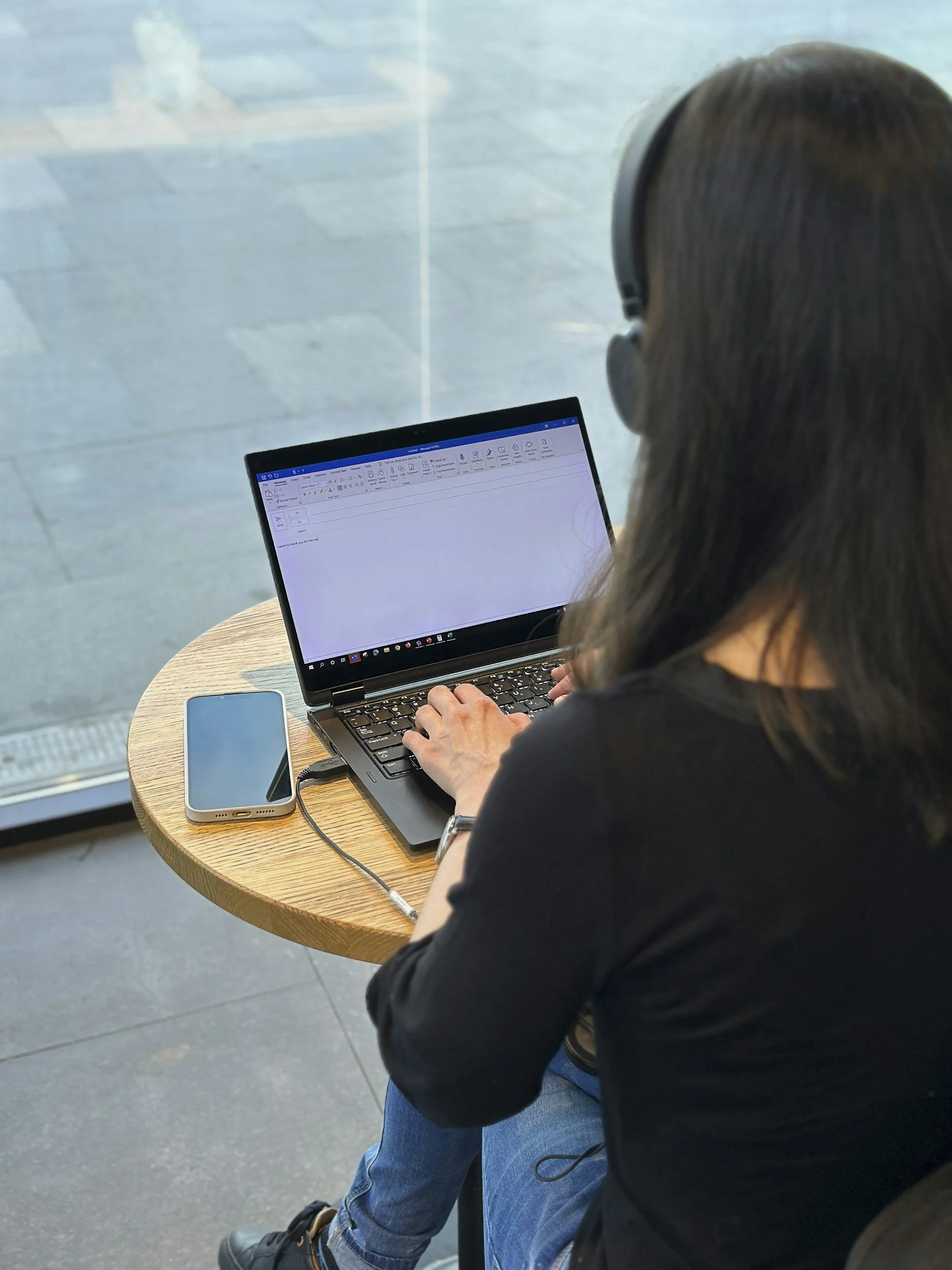 A person wearing headphones, sitting at a small, round wooden table, working on a laptop, with a smartphone nearby, in a bright indoor setting with a large window.