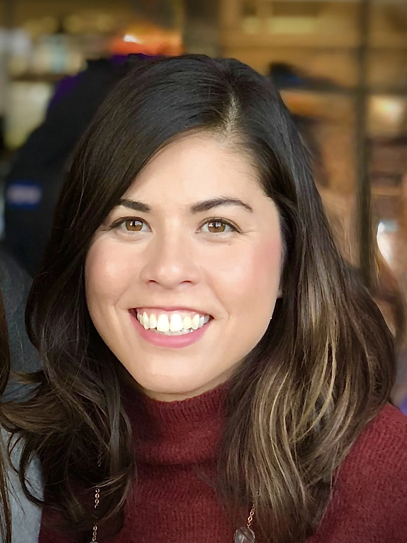 Close-up of a woman's face smiling, with long wavy brown hair, wearing a red sweater and a necklace, blurred background.