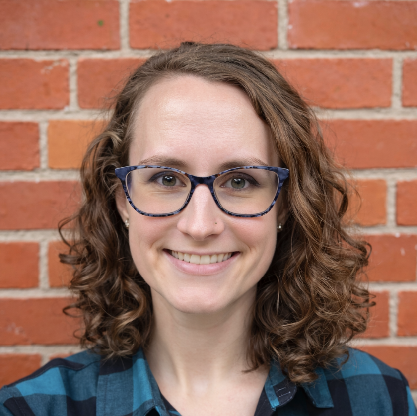 A woman with curly brown hair, wearing glasses and a blue checkered shirt, standing in front of a brick wall, smiling at the camera.