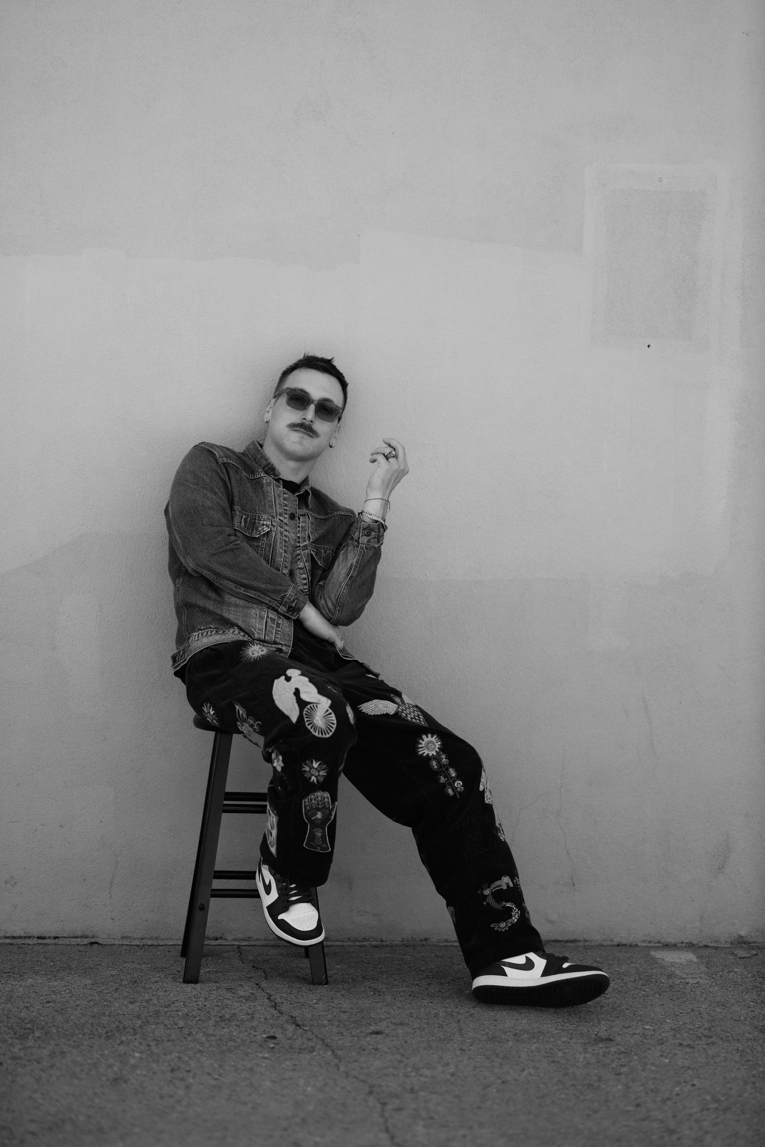 Black and white photo of a person sitting on a stool against a plain wall, wearing sunglasses, a denim jacket, and patterned pants with various designs, including flowers, hands, and symbols. The person has a mustache and short hair, and is holding a ring with their right hand near their face, with a relaxed posture and one leg crossed over the other.