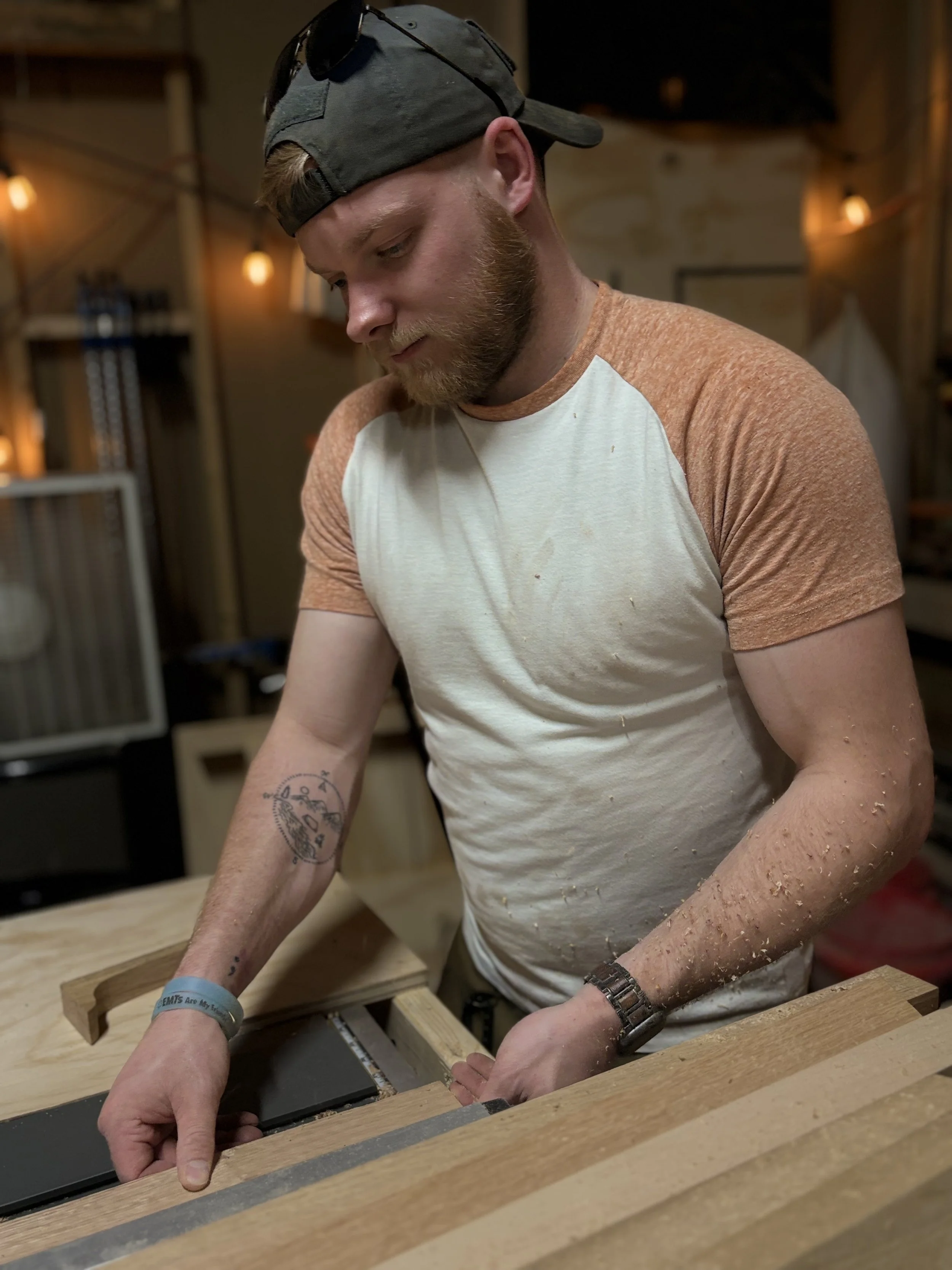 A man with a baseball cap, sunglasses, and a T-shirt working on a woodworking project in a workshop.