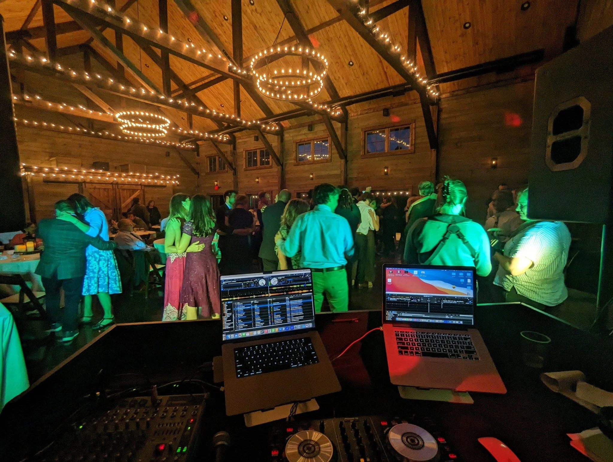 DJ or music artist's setup at an event with people dancing in a wooden loft with string lights and circular chandeliers.