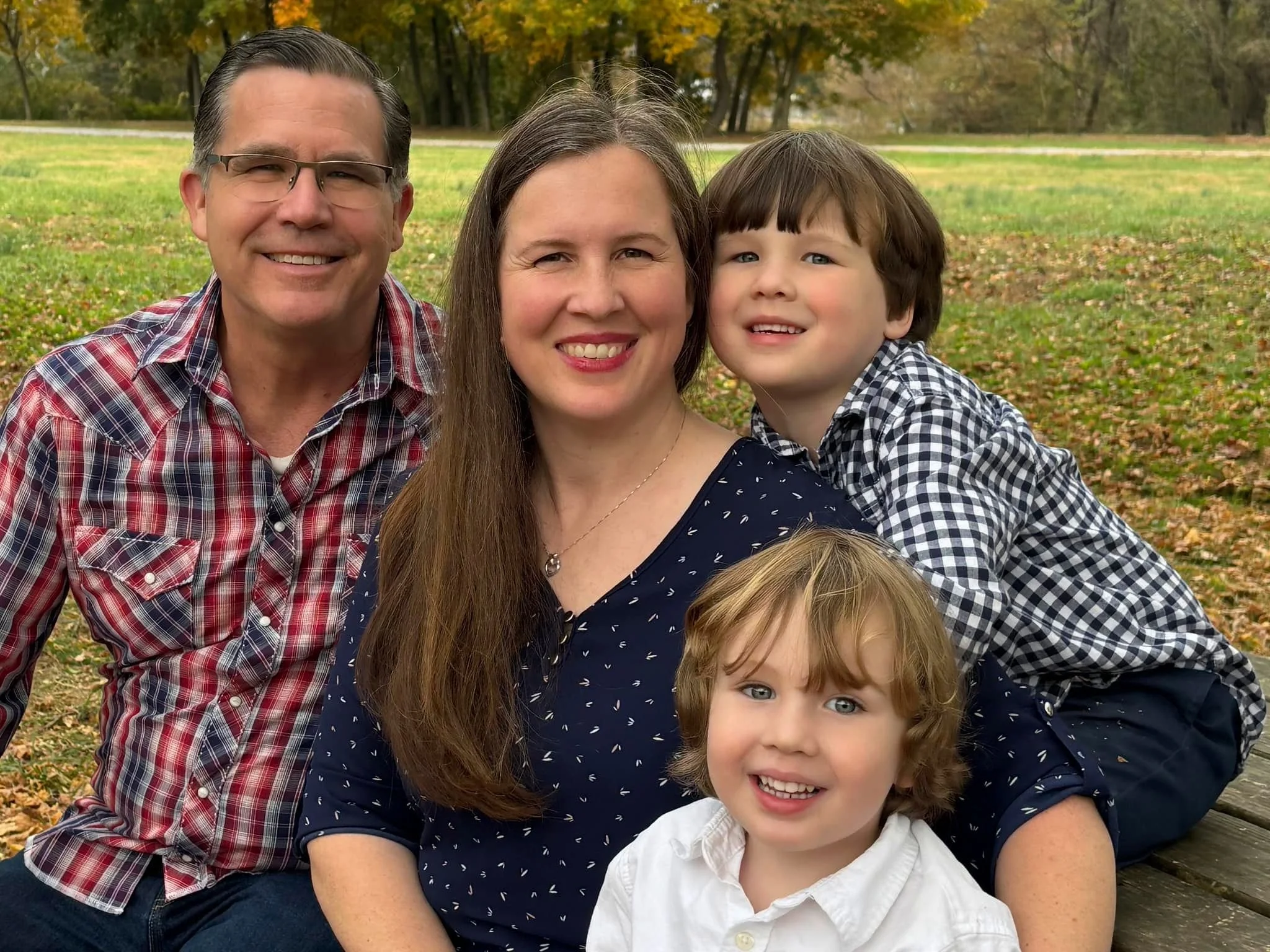A family of four taking a selfie outdoors in a park during fall. The background shows trees with autumn leaves.