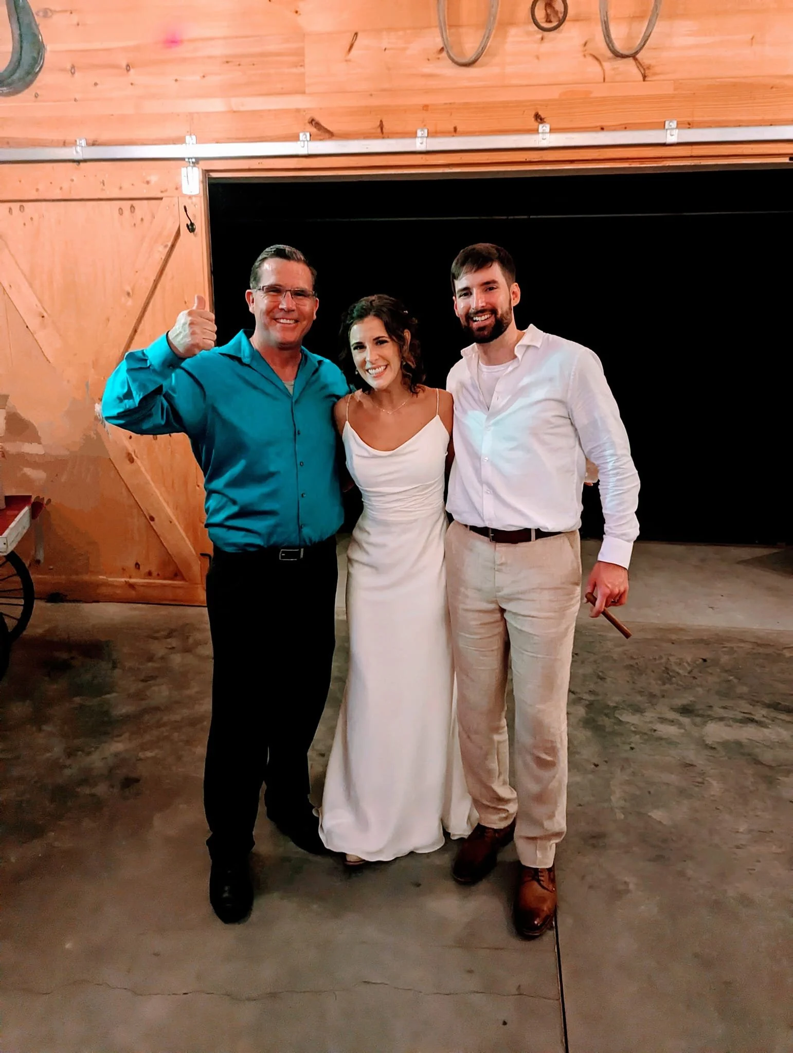 Three people standing together indoors, smiling at the camera. The person on the left is a man wearing glasses and a turquoise shirt, giving a thumbs-up. The middle person is a woman wearing a white dress, with curly dark hair. The person on the right is a man with a beard, wearing a white shirt and beige pants. They are in front of a black background with a wooden wall and some bicycles hanging above them.
