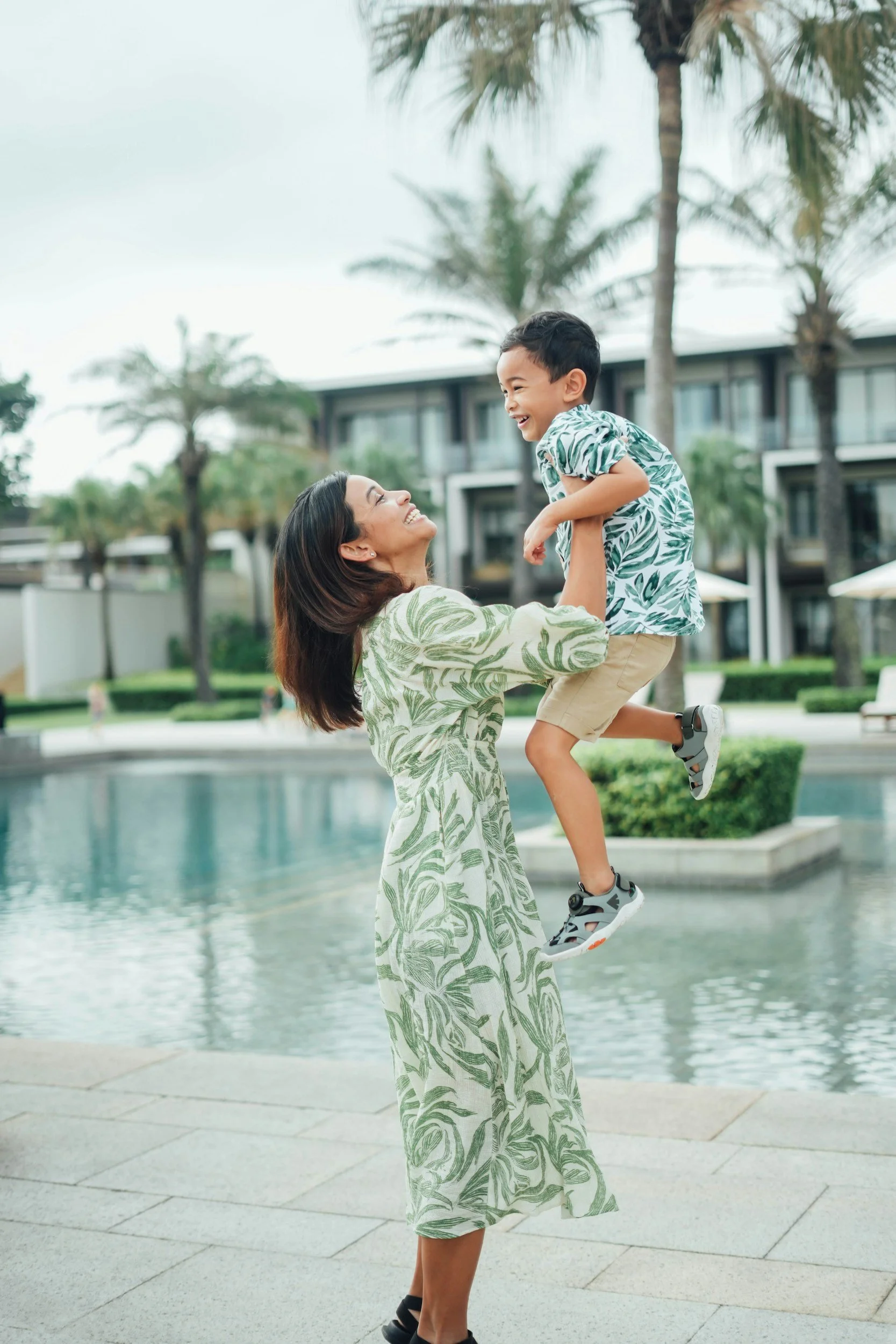 A woman in a green leafy patterned dress lifting a smiling young boy in a matching shirt in an outdoor setting near a pool and palm trees, with modern buildings in the background.