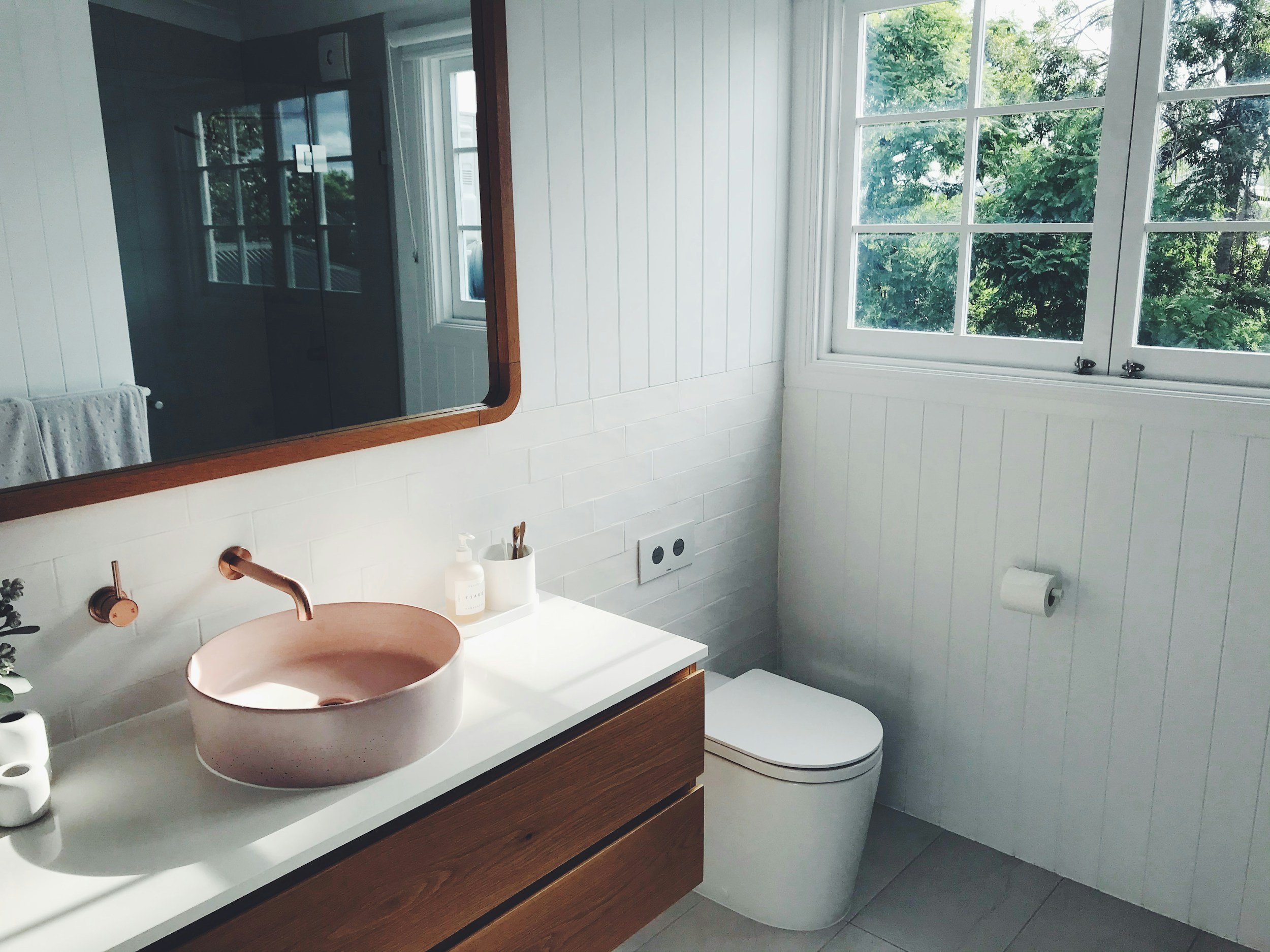 A modern bathroom with white walls, a large window with a view of green trees, a wooden vanity with a round stone sink, a soap dispenser, toothbrush holder, and a towel, and a toilet. A mirror is mounted above the sink, and there is a black cabinet with reflective doors on the wall.