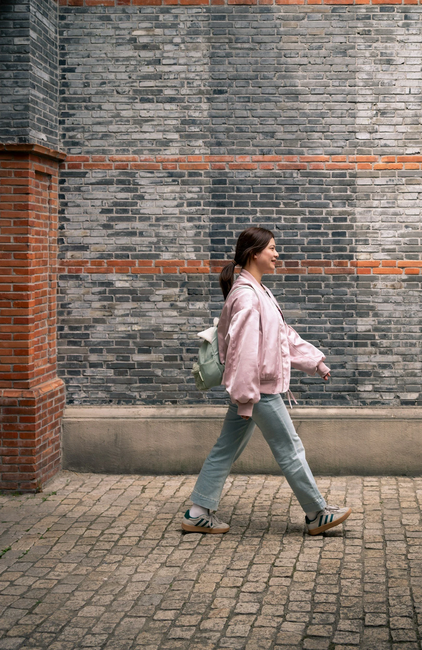 A young woman walking on a cobblestone street next to a brick wall, wearing a pink satin jacket, gray pants, and sneakers, with a green backpack.
