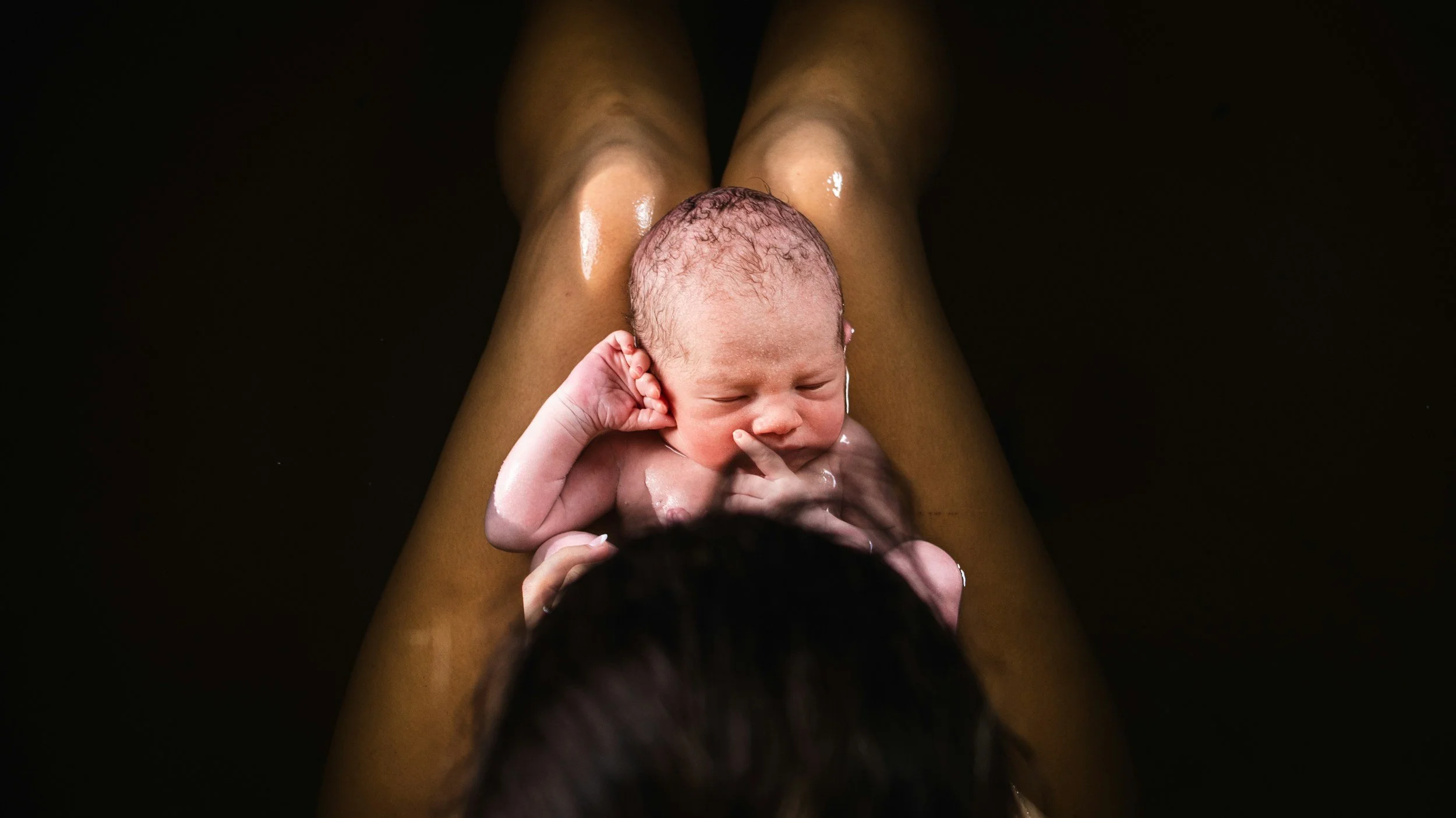 A newborn baby being held closely by a person, with the person's legs visible and the baby's head resting on their leg.