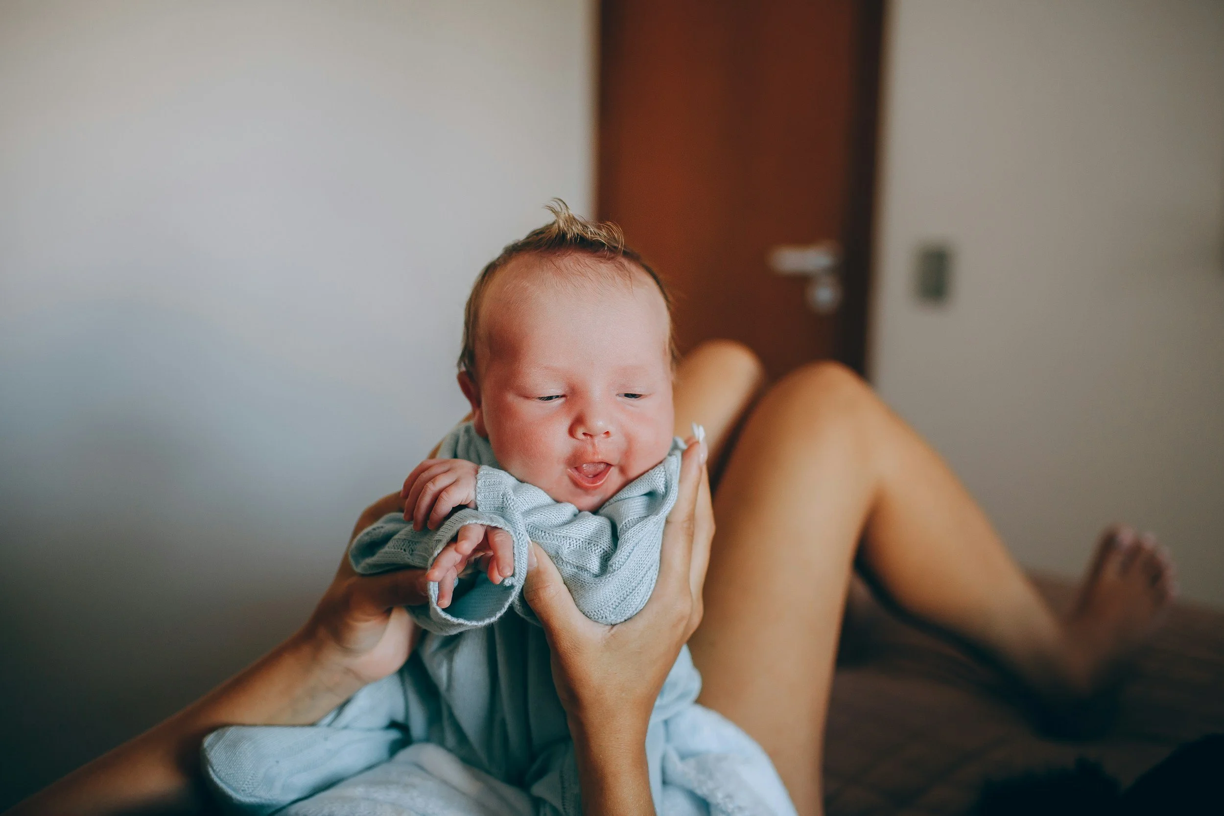 A baby with reddish hair being held up by someone with a bare arm in a room with a wooden door in the background.