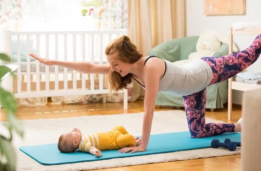 A woman exercising on a blue yoga mat with a baby lying on its back nearby, in a well-lit living room.