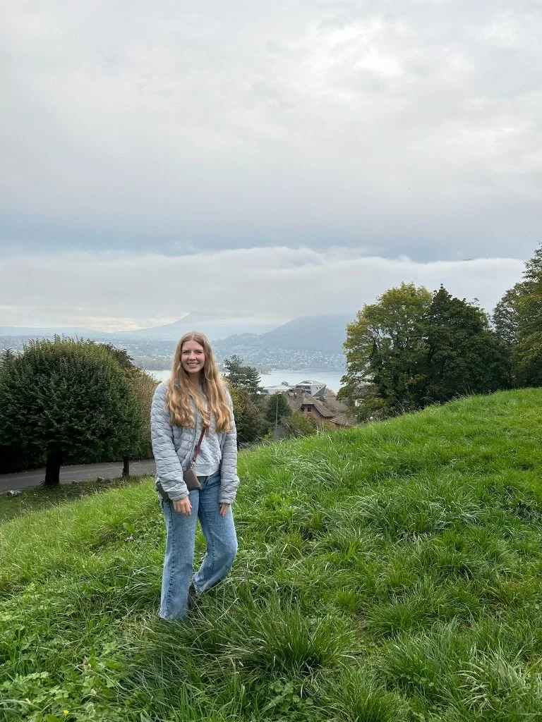 A woman standing on a grassy hillside with trees, a body of water, and mountains in the background under a cloudy sky.