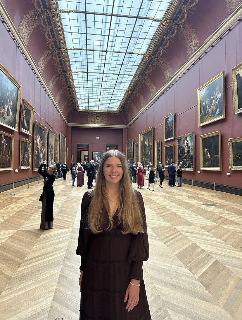 A woman with long hair in a dark dress standing in an art gallery with ornate red walls, gold picture frames, and large paintings. The gallery has a high glass ceiling, and other visitors are seen in the background.