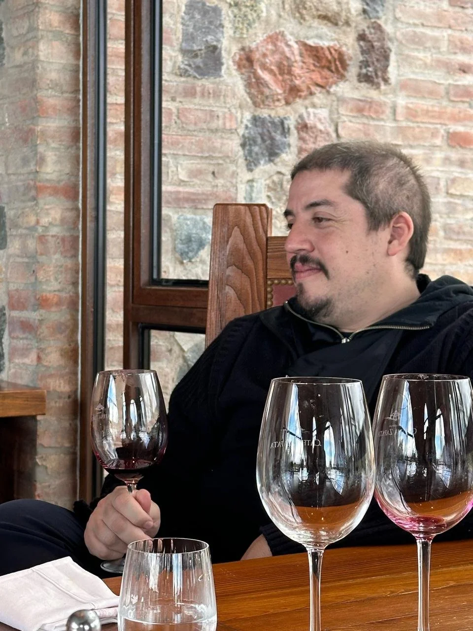 Hombre sentado en una mesa con varias copas de vino y una copa en mano, en un ambiente con paredes de piedra.