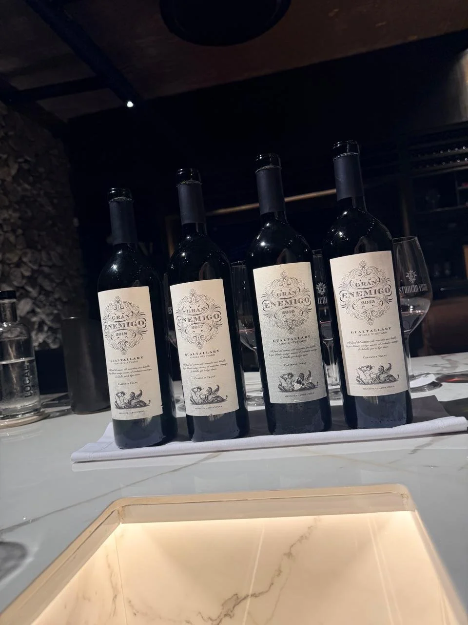 Cuatro botellas de vino tinto sobre una mesa blanca en un ambiente de restaurante o bodega.