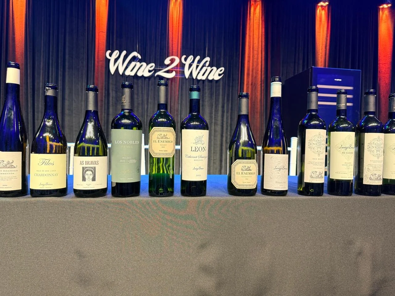 Filas de botellas de vino sobre una mesa, fondo con cortinas azules y un letrero que dice 'Wine 2 Wine' en letras blancas y rojas.