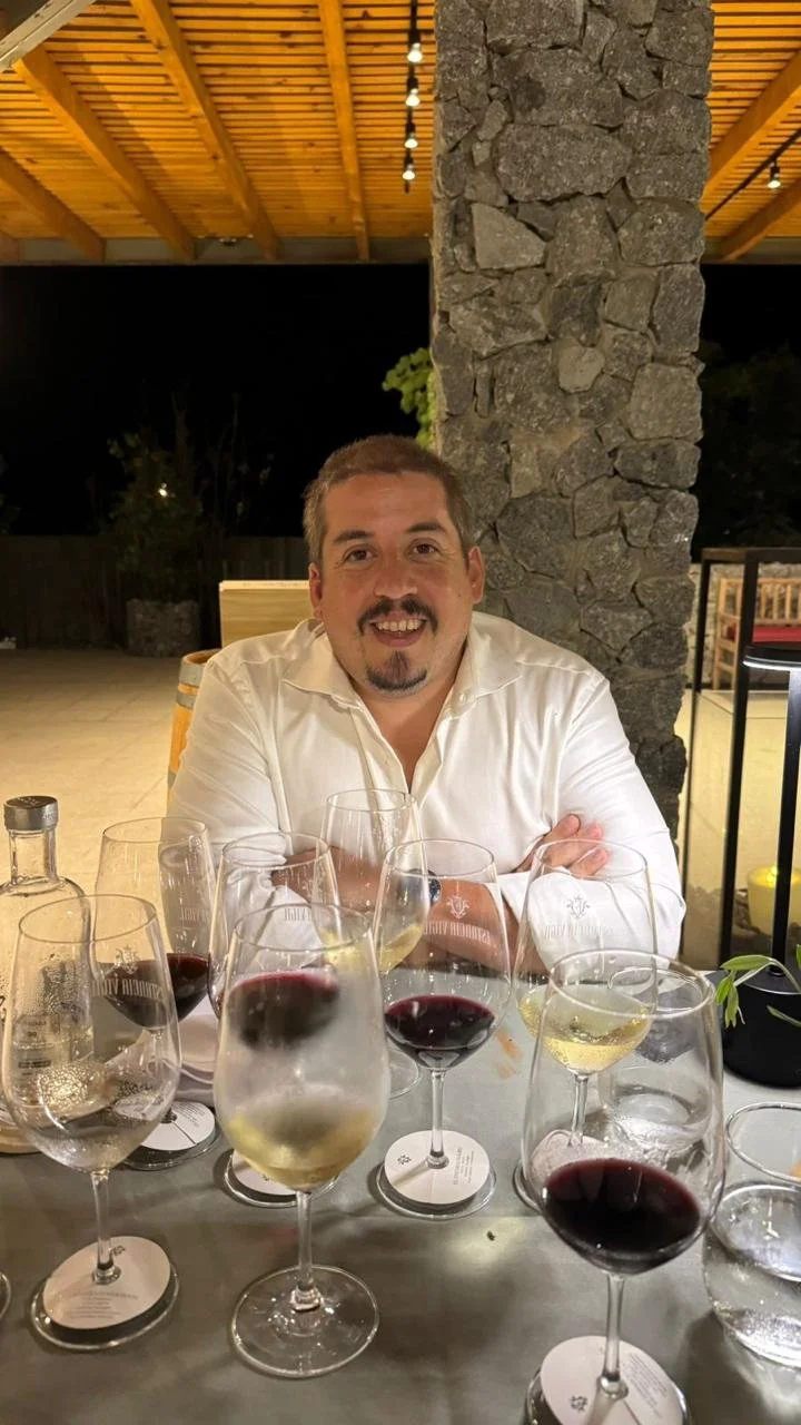 Hombre sonriendo sentado en una mesa con varias copas de vino tinto y vino blanco, en un ambiente de restaurante en la noche.