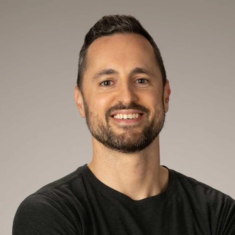 Smiling man with short dark hair, beard, and mustache, wearing a black t-shirt against a plain background.
