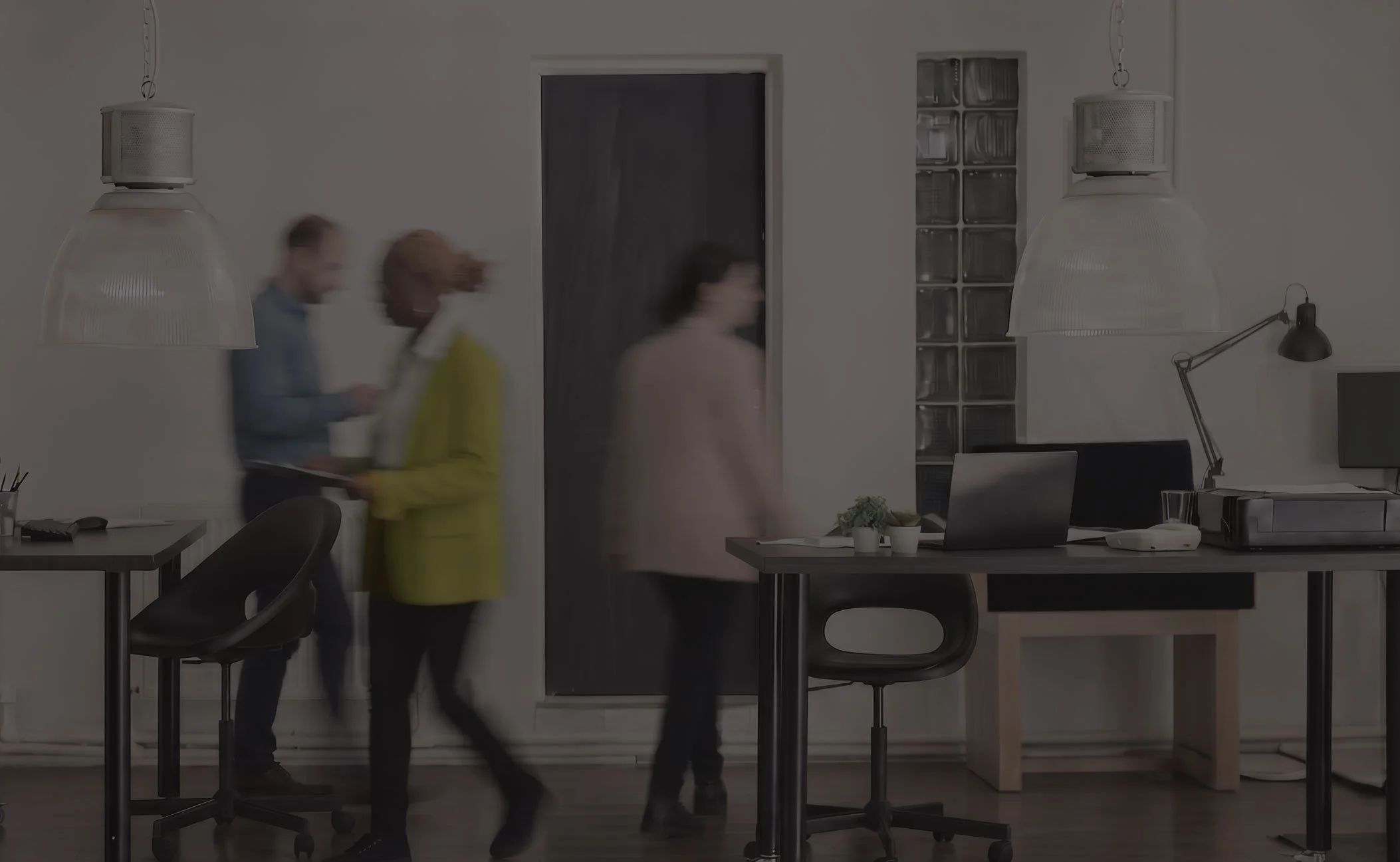 An office with two people walking past a desk, blurred due to motion, with modern lighting and furniture, and a window with glass blocks.