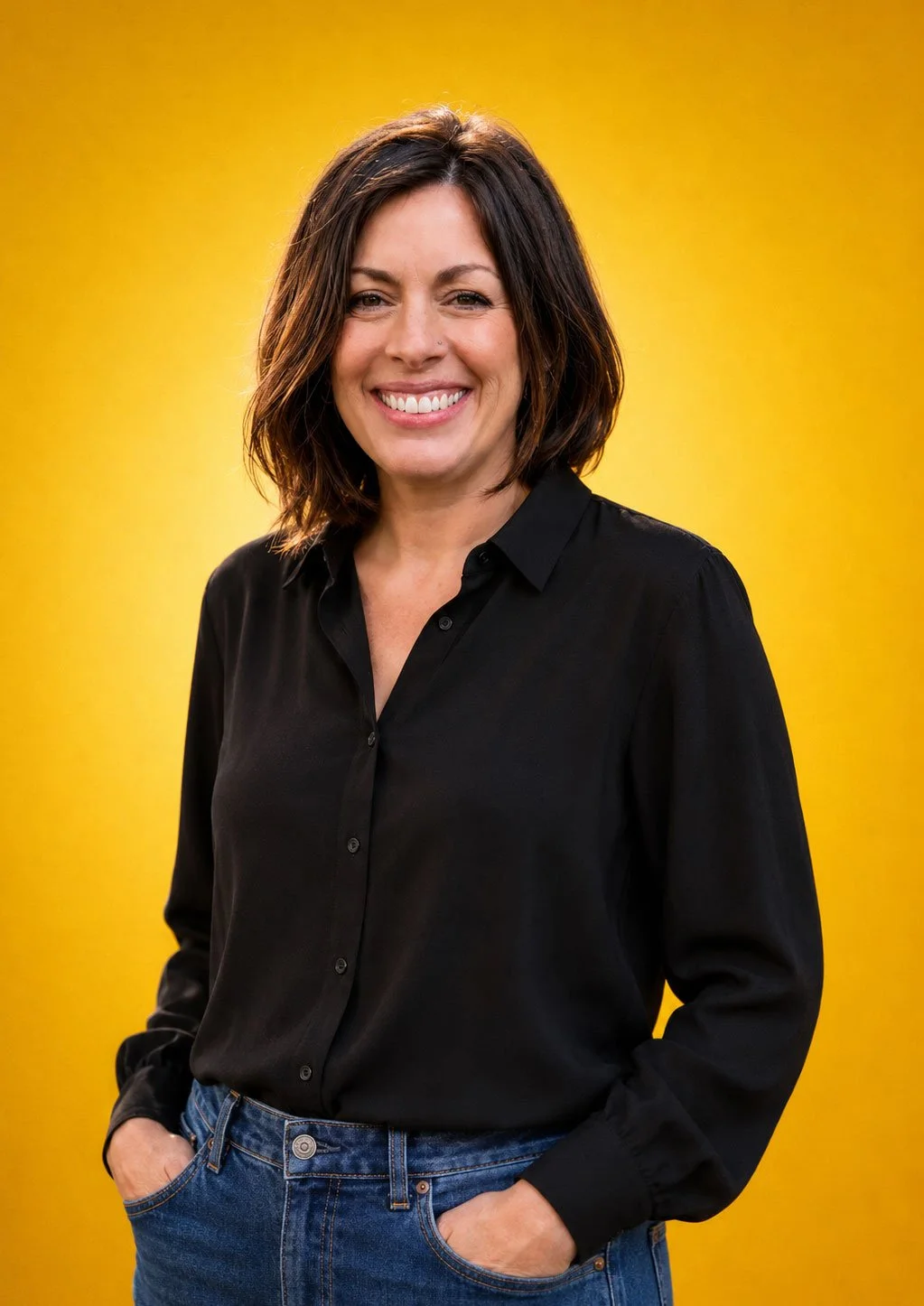 A woman with short dark hair, wearing a black button-up shirt and blue jeans, smiling with her hands in her pockets against a yellow background.