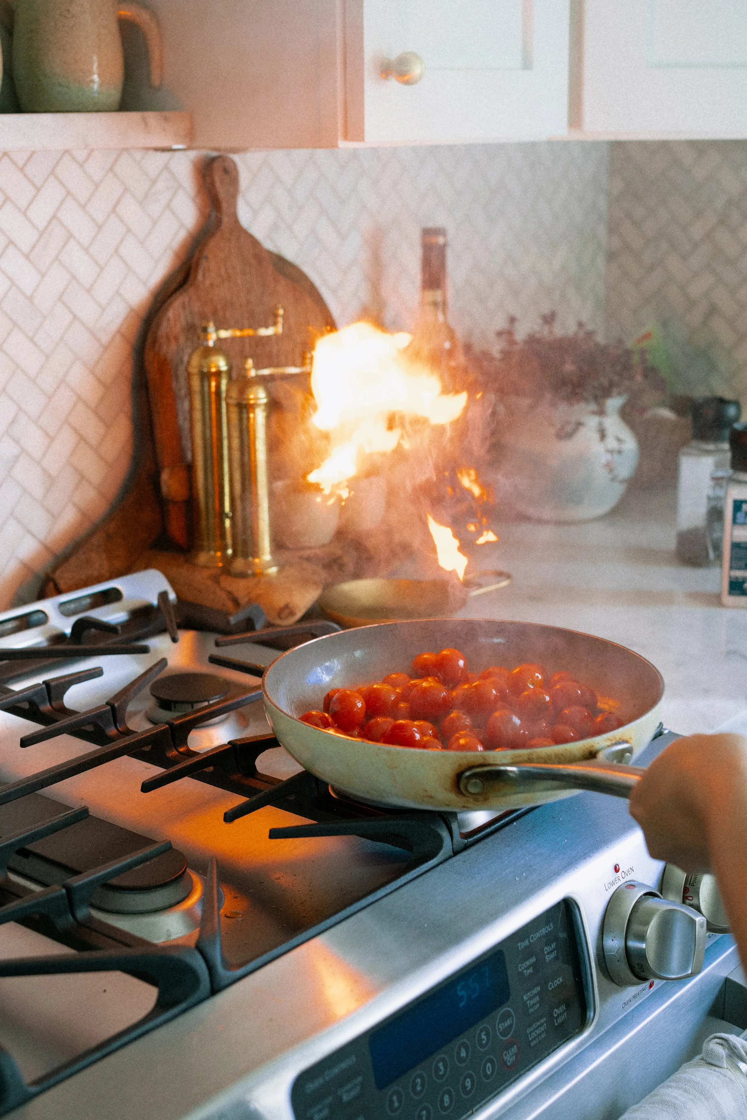 A stove with a pan of cherry tomatoes and a small flame coming from the back of the stove, with kitchen cabinets and decorative items on the counter in the background.