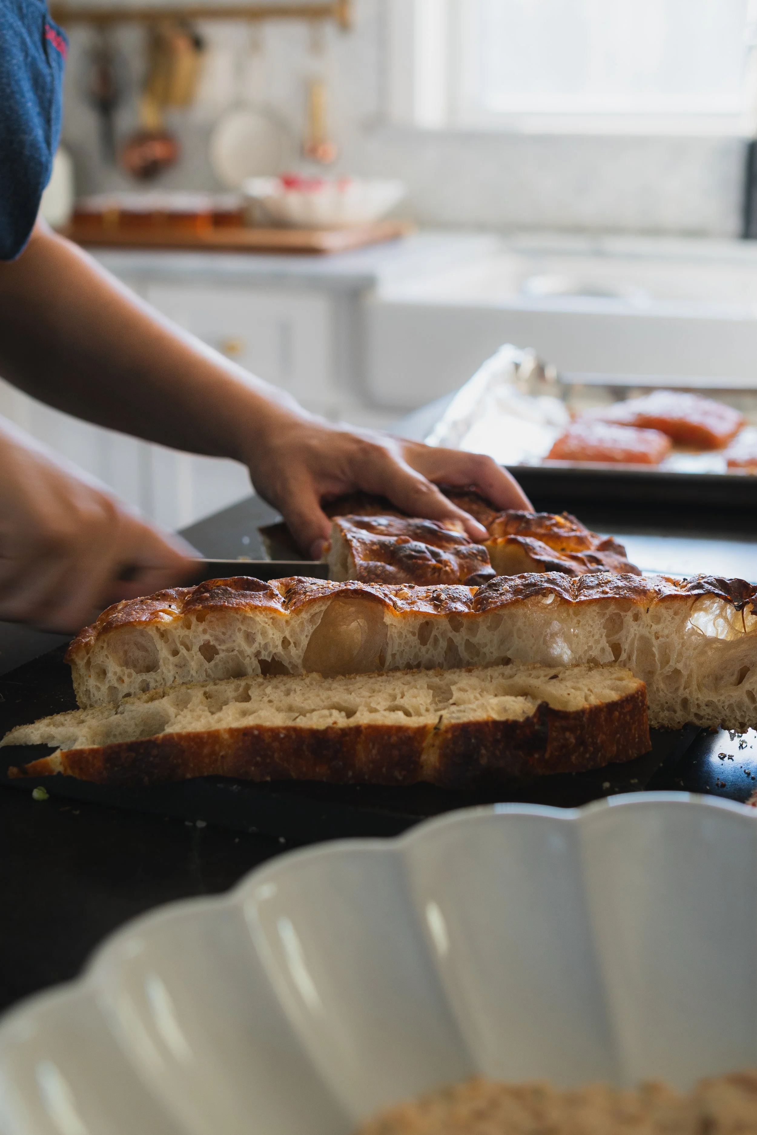 Person slicing freshly baked loaf of bread in a kitchen with a tray of salmon or cured meat in the background.