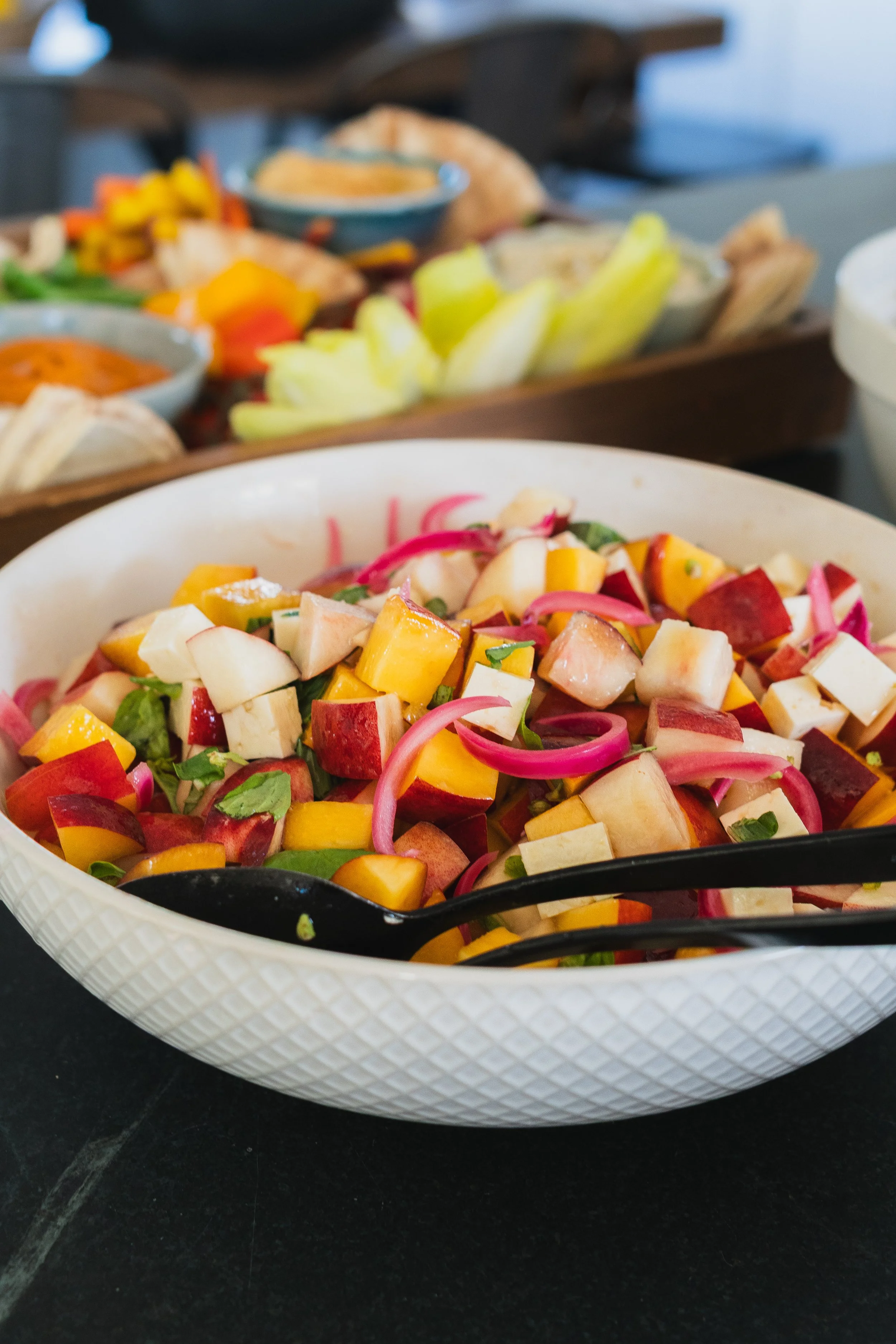 A white bowl filled with a colorful chopped fruit salad. The fruit includes pieces of apple, mango, and possibly other fruits, garnished with herbs. A black spoon rests inside the bowl. In the background, there is a wooden serving tray with various b